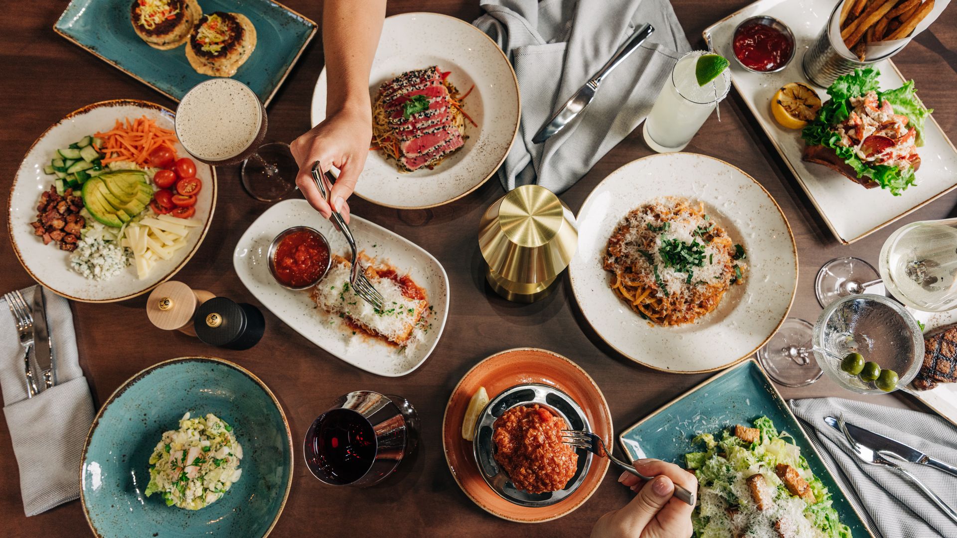 Top-down view of a wooden table filled with a family-style feast: salad with avocado, tomatoes and cucumber; pasta; sesame meat slices; tacos; fries; sauces; a lime cocktail; and hands serving food.