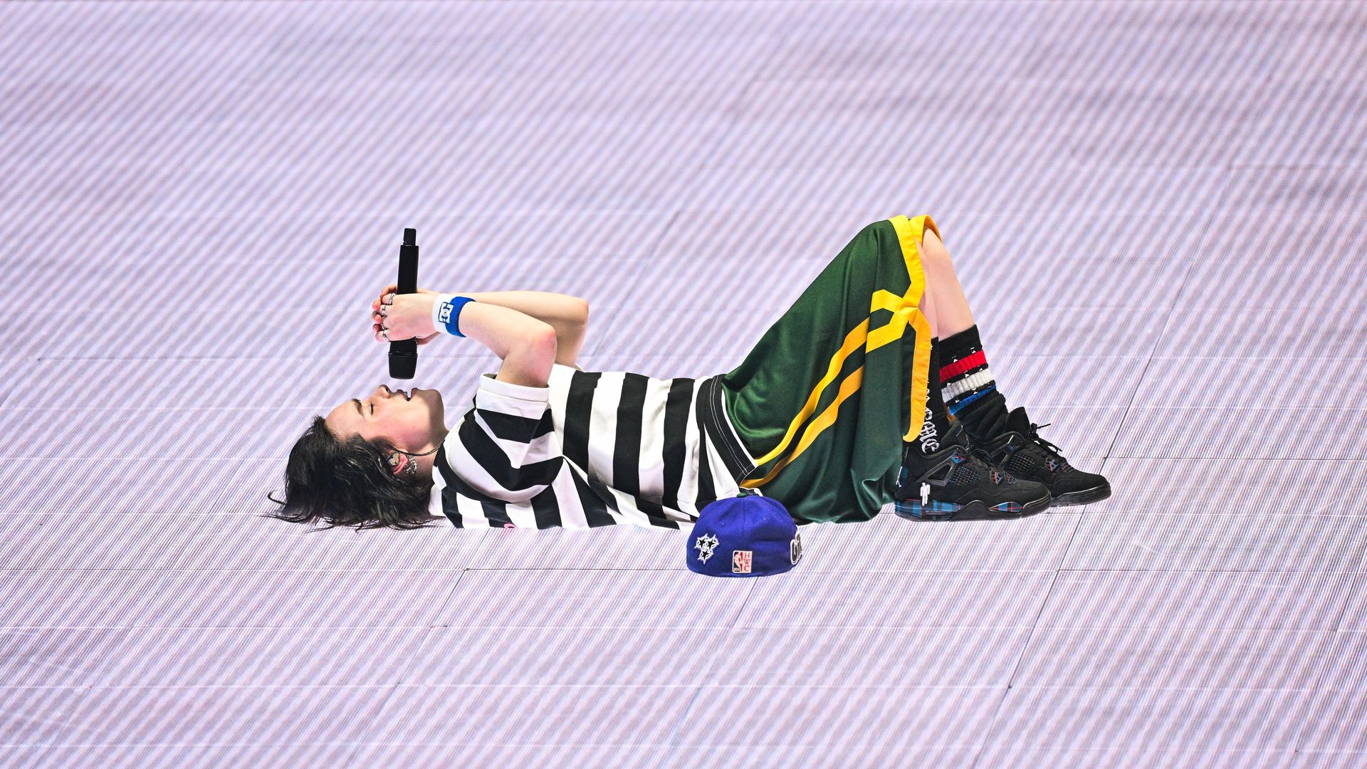 Billie Eilish sings into a microphone while laying flat on her back on a video panel that looks like gray-white lines.