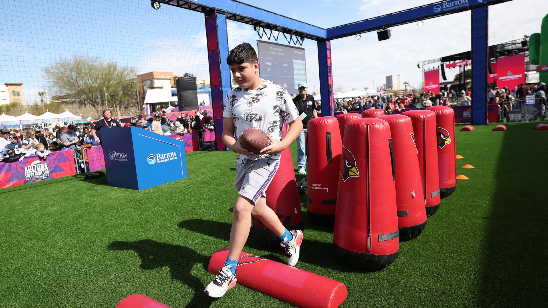 A young boy holds a football as he jumps over red padded obstacles.