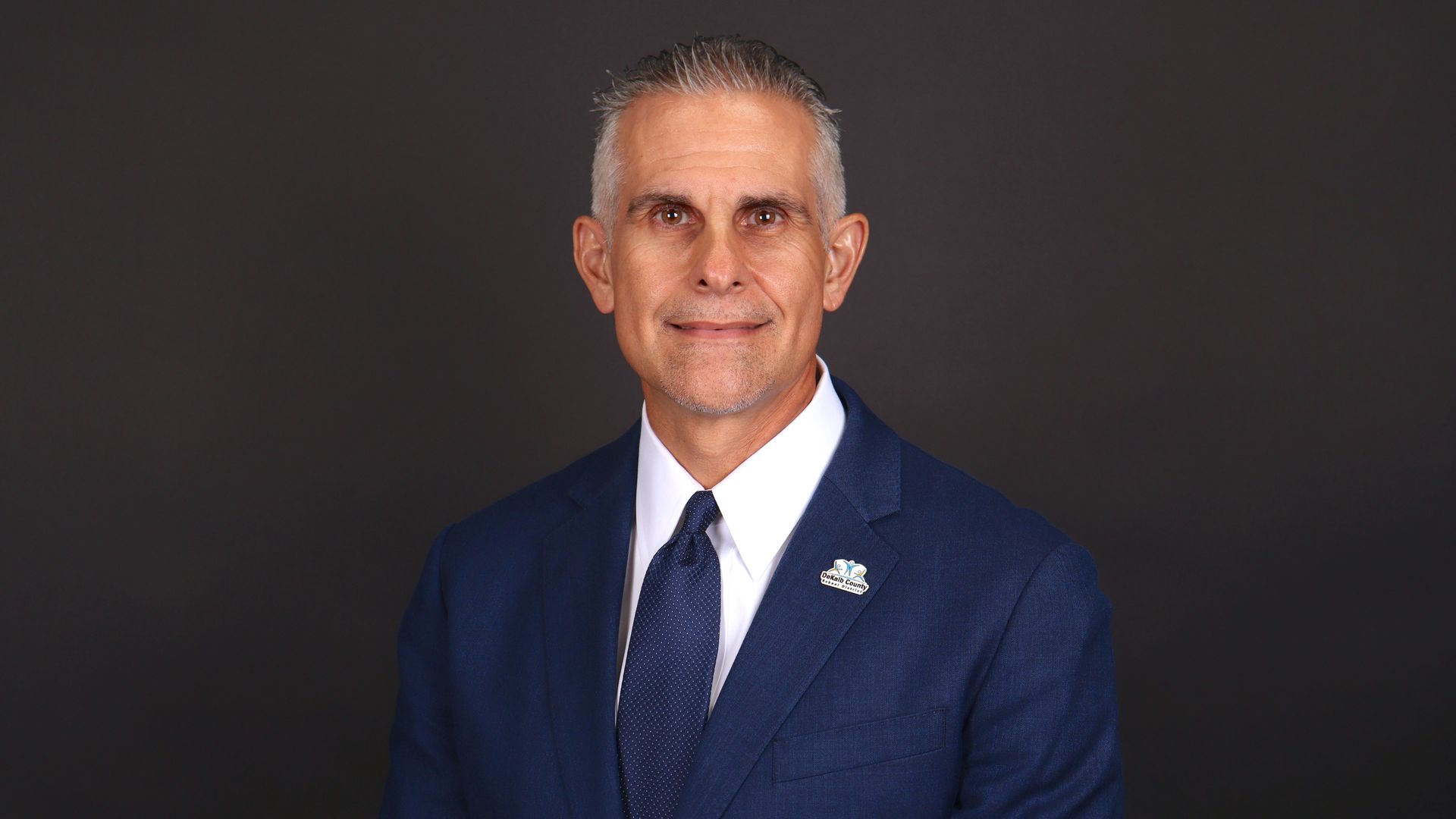 Portrait of a middle-aged man with short gray hair wearing a navy suit, white shirt, and dark blue tie, standing against a dark gray background; a small lapel pin is visible on the left.