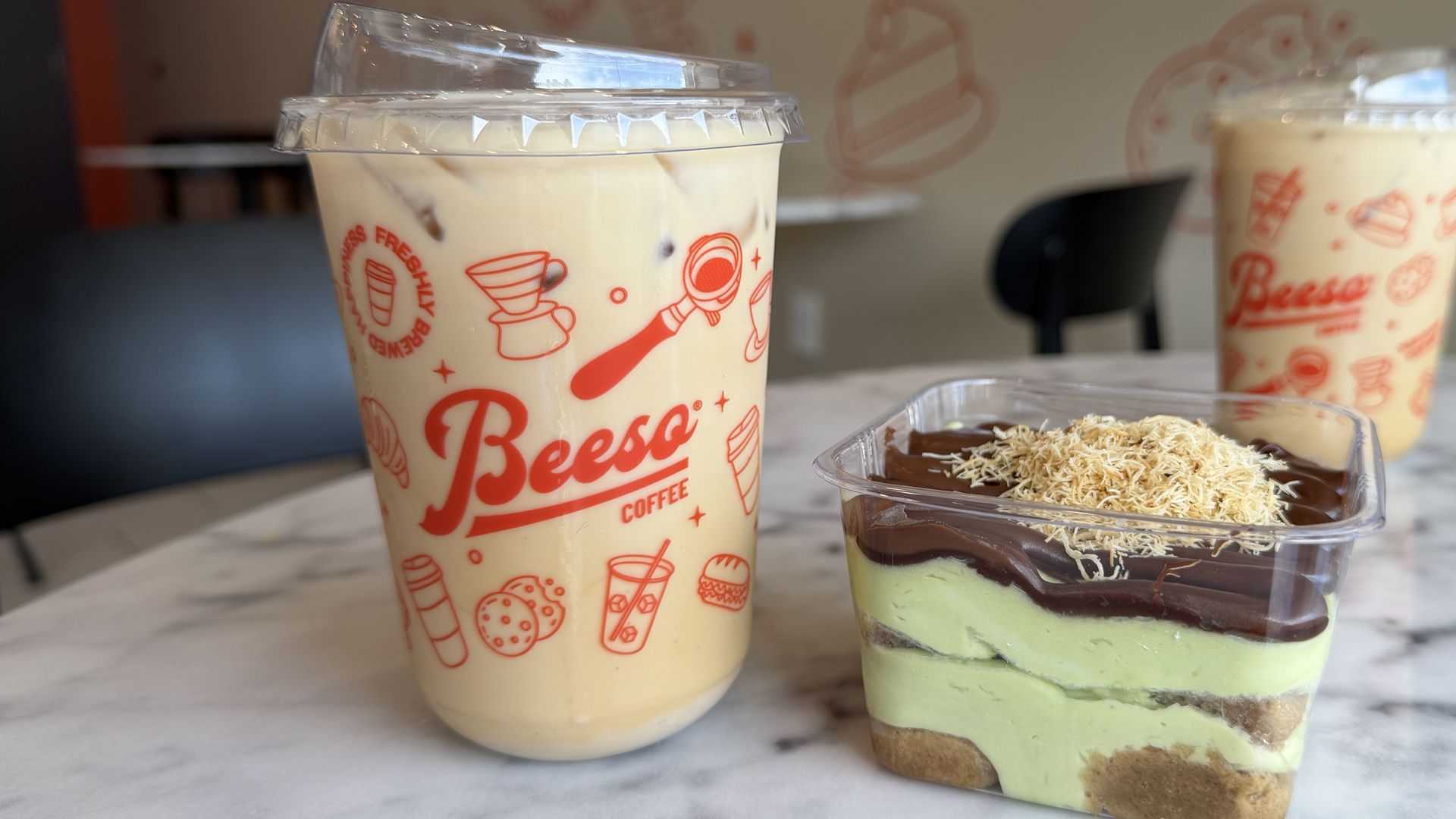 Two iced coffee cups with red graphics and plastic lids on a marble table; a clear container holds a layered pistachio dessert topped with chocolate and coconut shavings in the foreground.