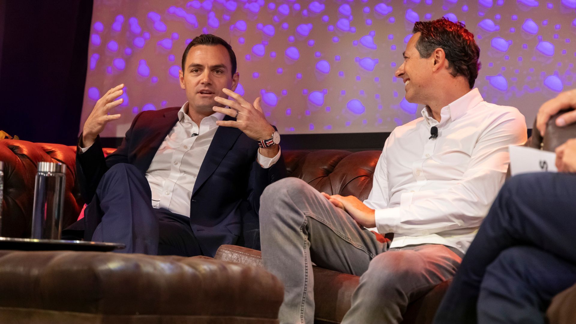 Two men on brown leather couches in a discussion; one in a navy suit and white shirt, the other in a white shirt and gray pants, with a backdrop of purple light patterns.