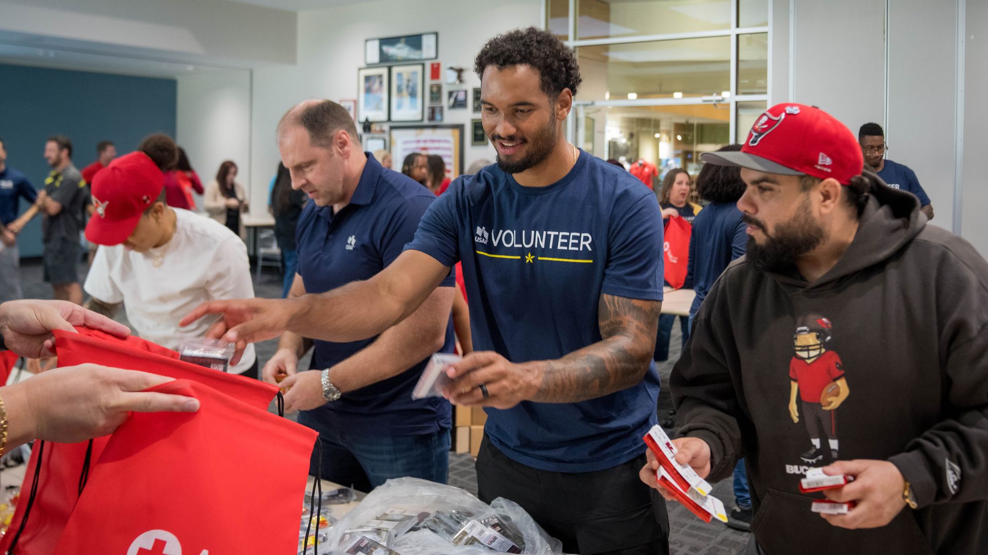 Inside a community center, volunteers hand out supplies. A man in a blue "VOLUNTEER" shirt smiles as others reach for red American Red Cross bags, with more people in the background.