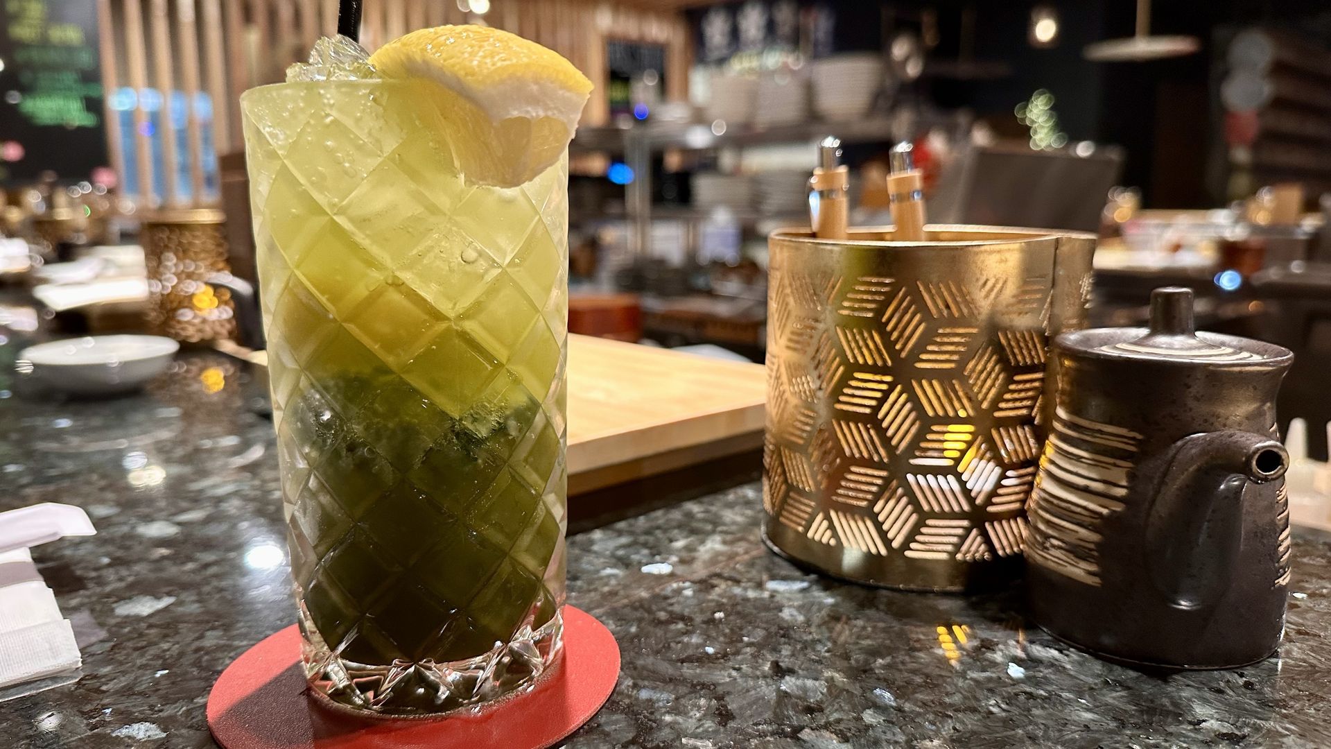 Close-up of a matcha iced drink in a textured glass with a lemon wedge on top, placed on a red coaster on a dark marble counter.