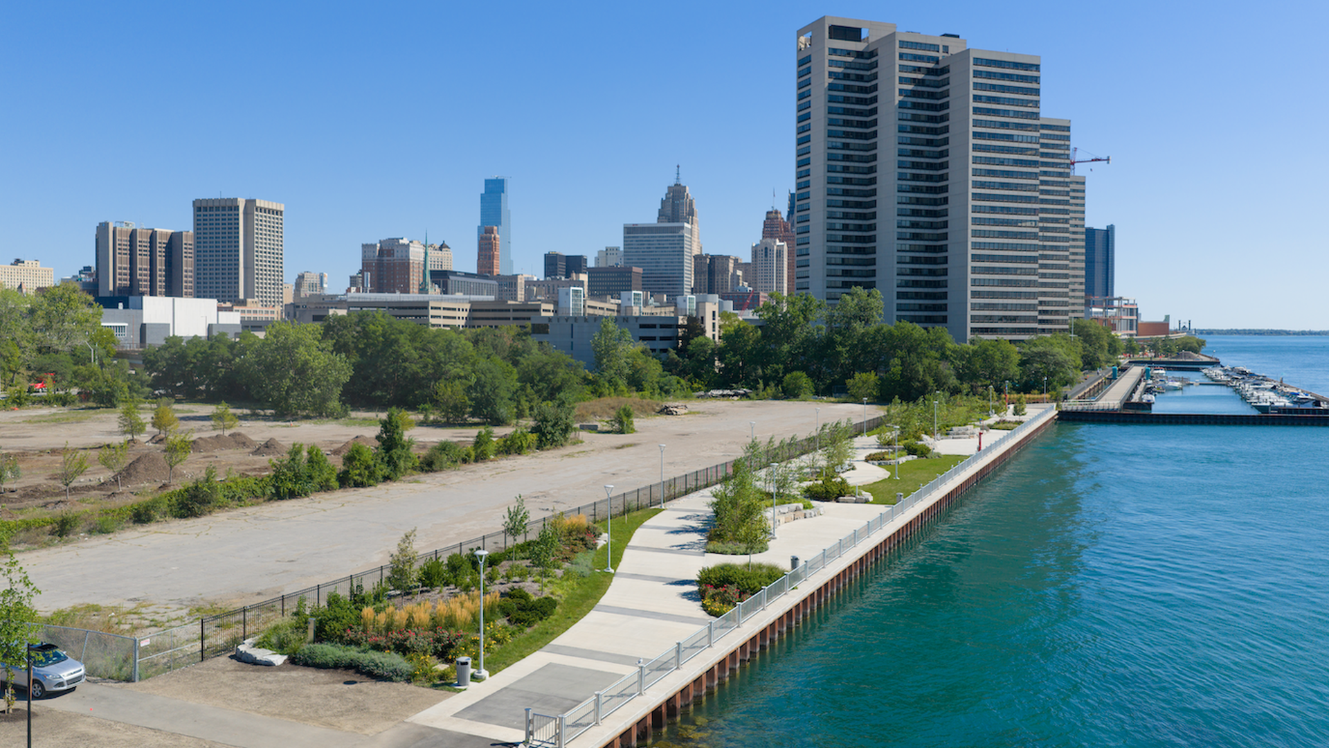 Waterfront promenade with green trees and plants along the river, high rise buildings. 