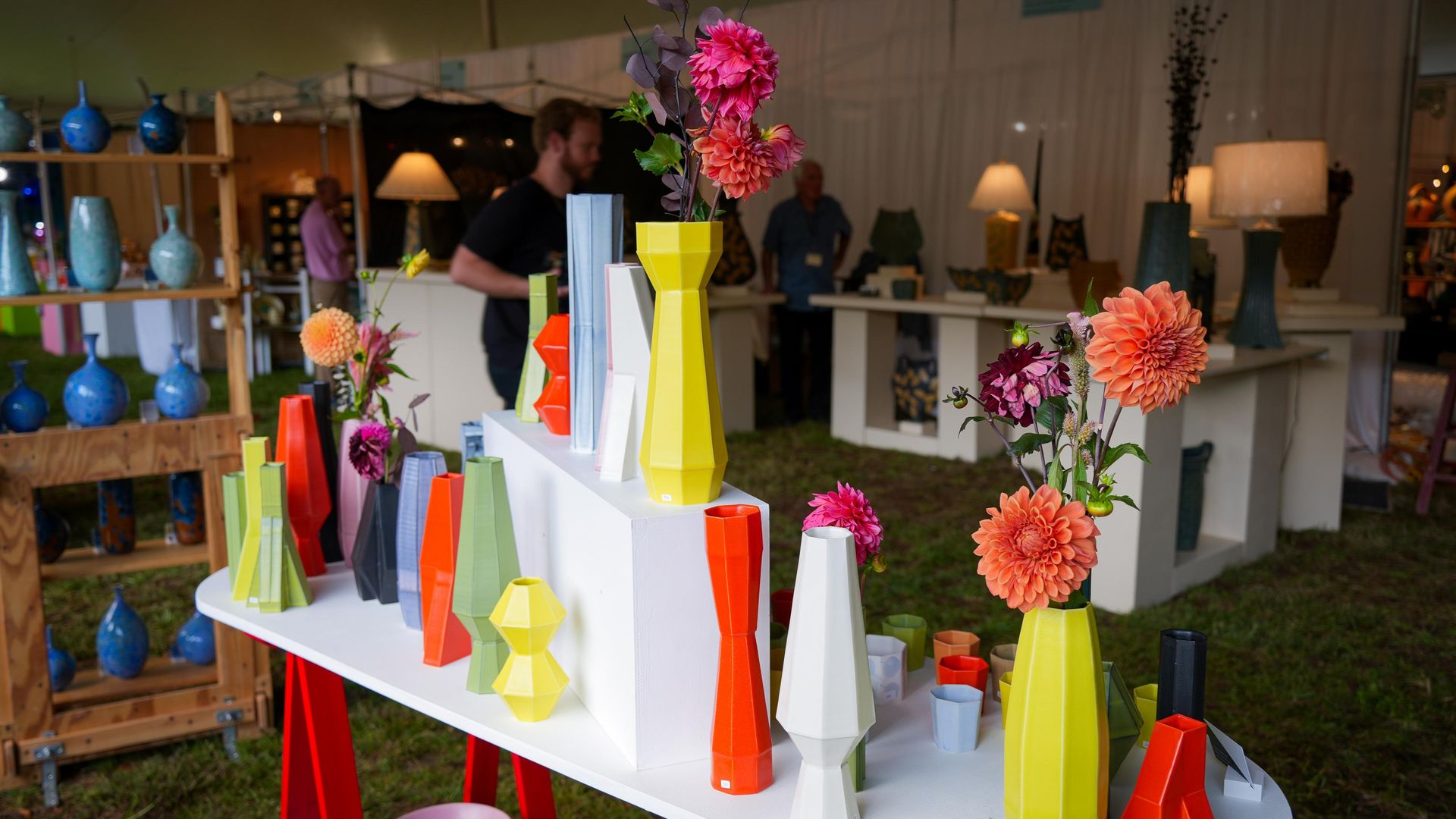 Colorful geometric vases in yellow, orange, white, green, and gray displayed on white tables with pink and purple flowers, set inside a tent with people and pottery in the background.