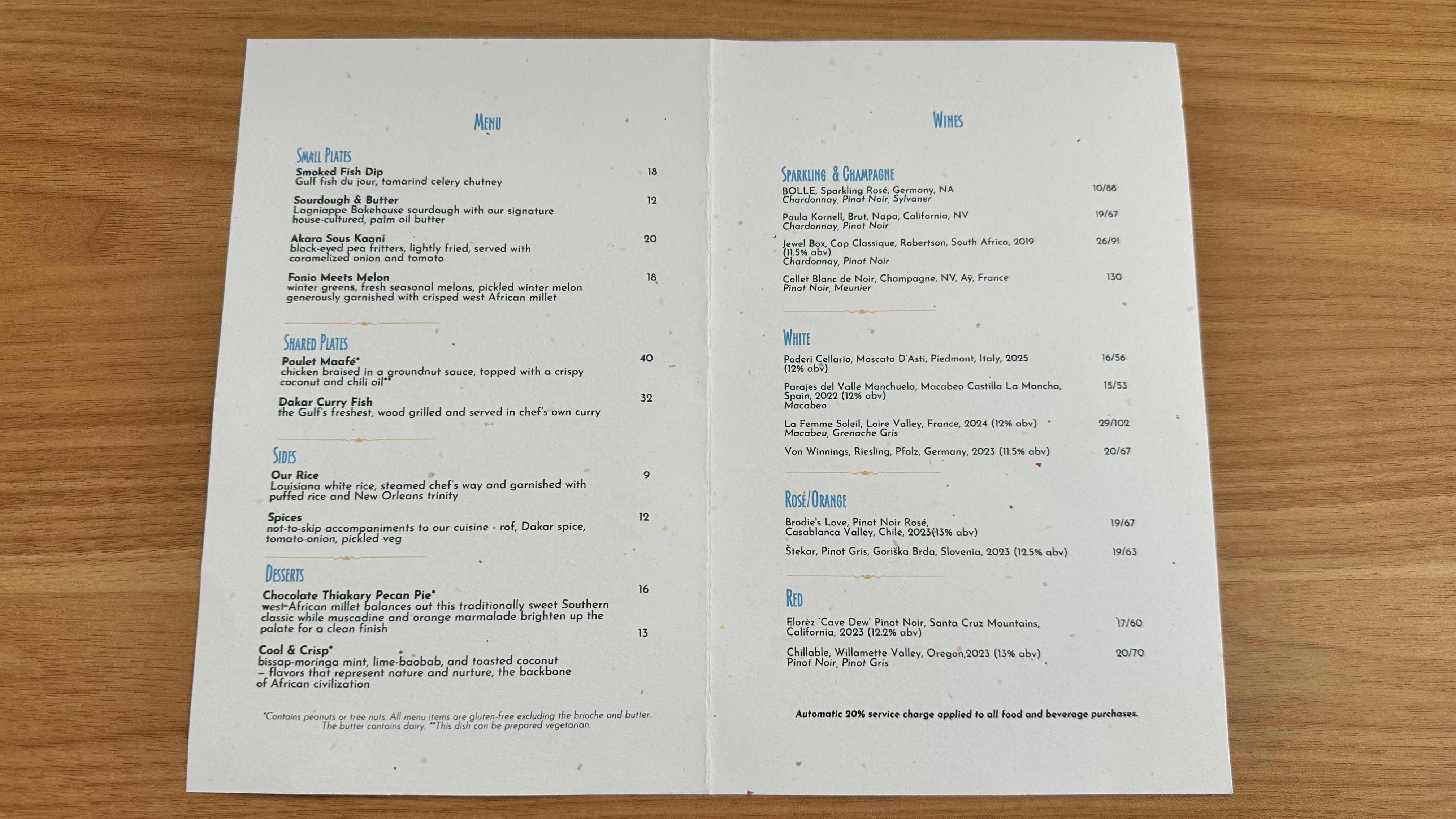 Open, two-page restaurant menu on a wooden table. Left page shows blue "Menu" heading with sections and dish names; right page titled "Wines" with lists and prices. Light speckled off-white paper.