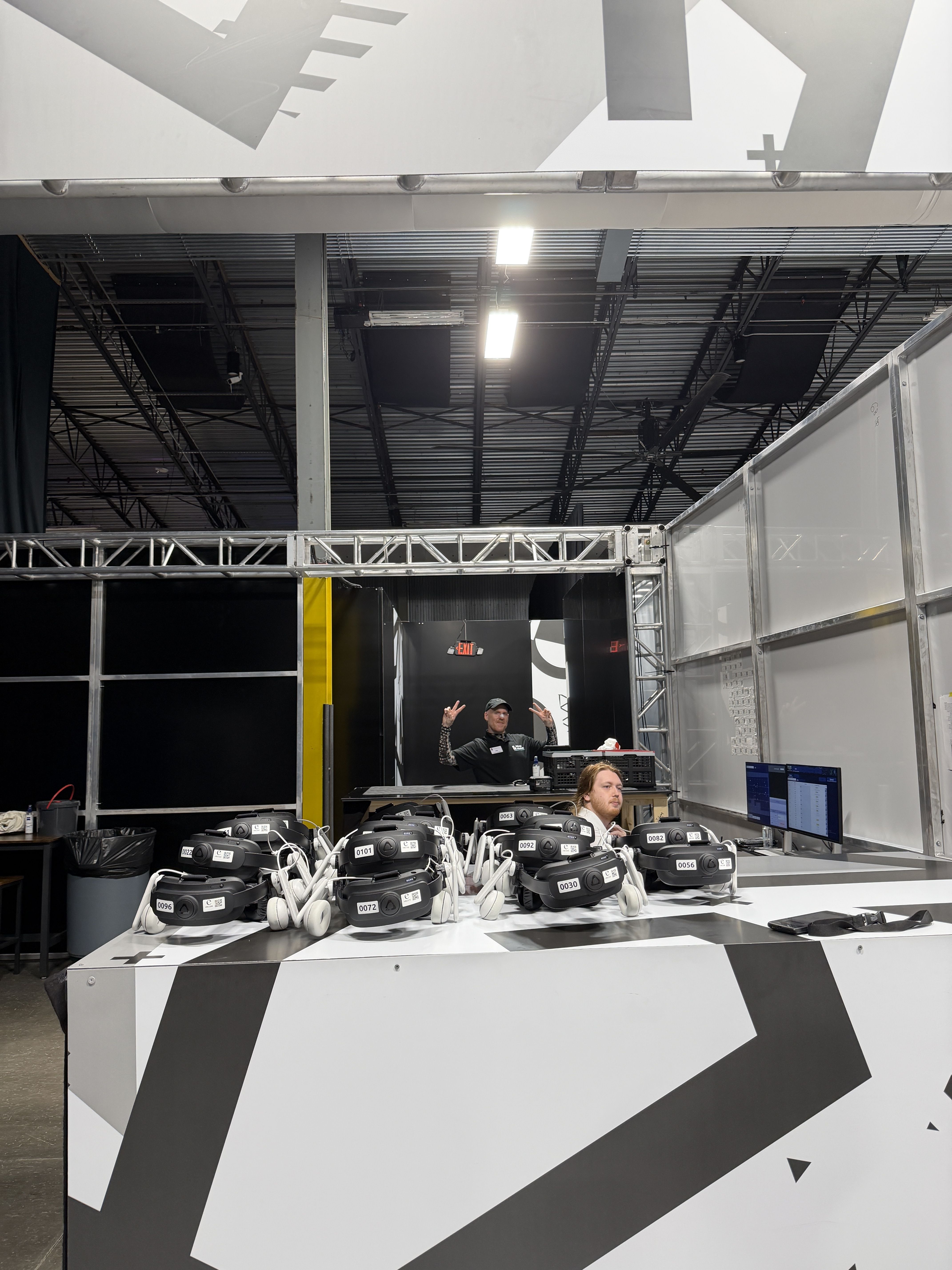 Trade show booth with a row of numbered white VR headsets on a counter; two staffers stand behind a metal truss structure under a gray geometric backdrop and bright ceiling lights.