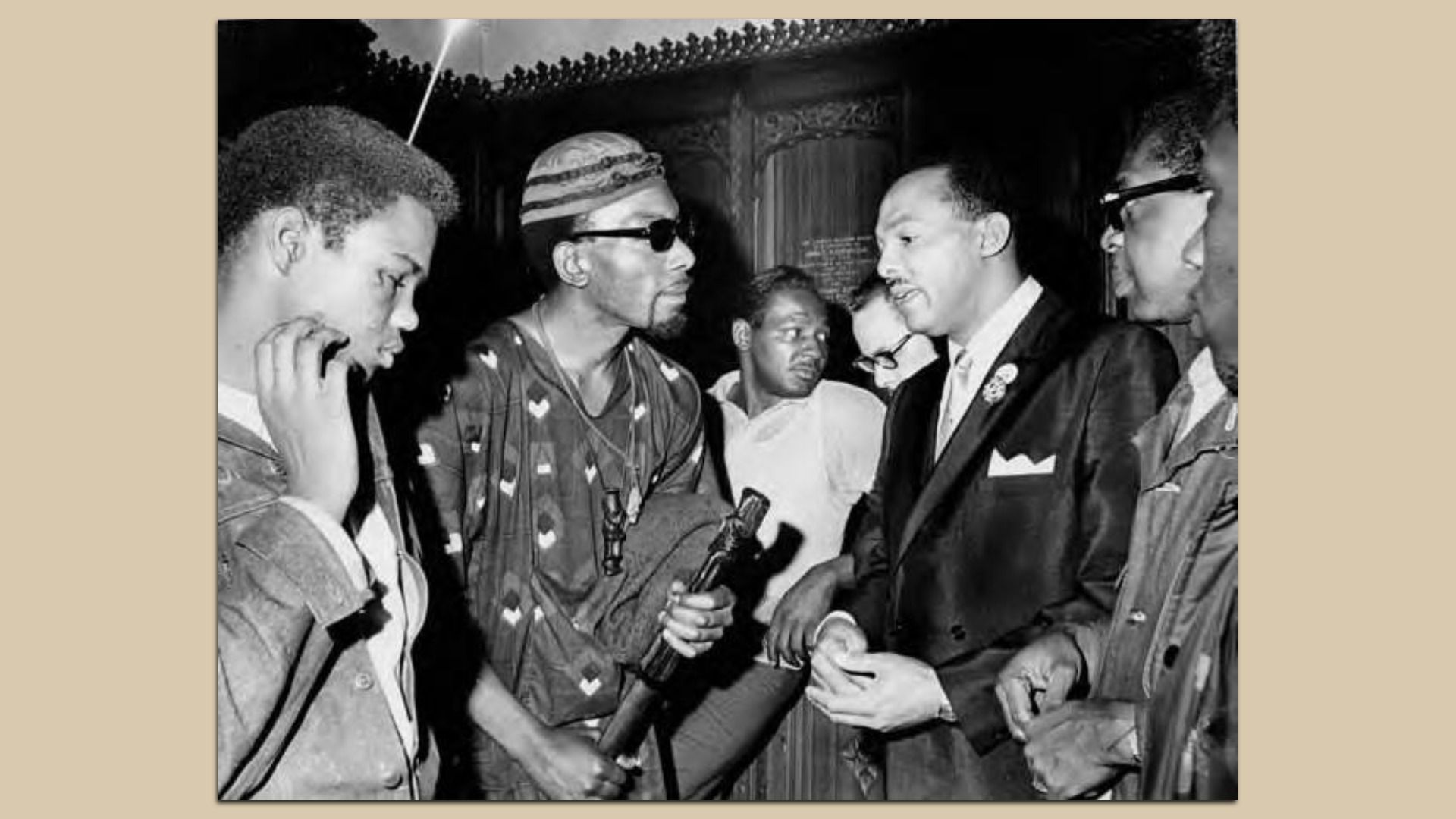 Black-and-white photo of a group of men in a street setting; two in suits exchange a handshake while others in casual wear gather around, one man wearing a striped bandana and sunglasses holds a microphone.