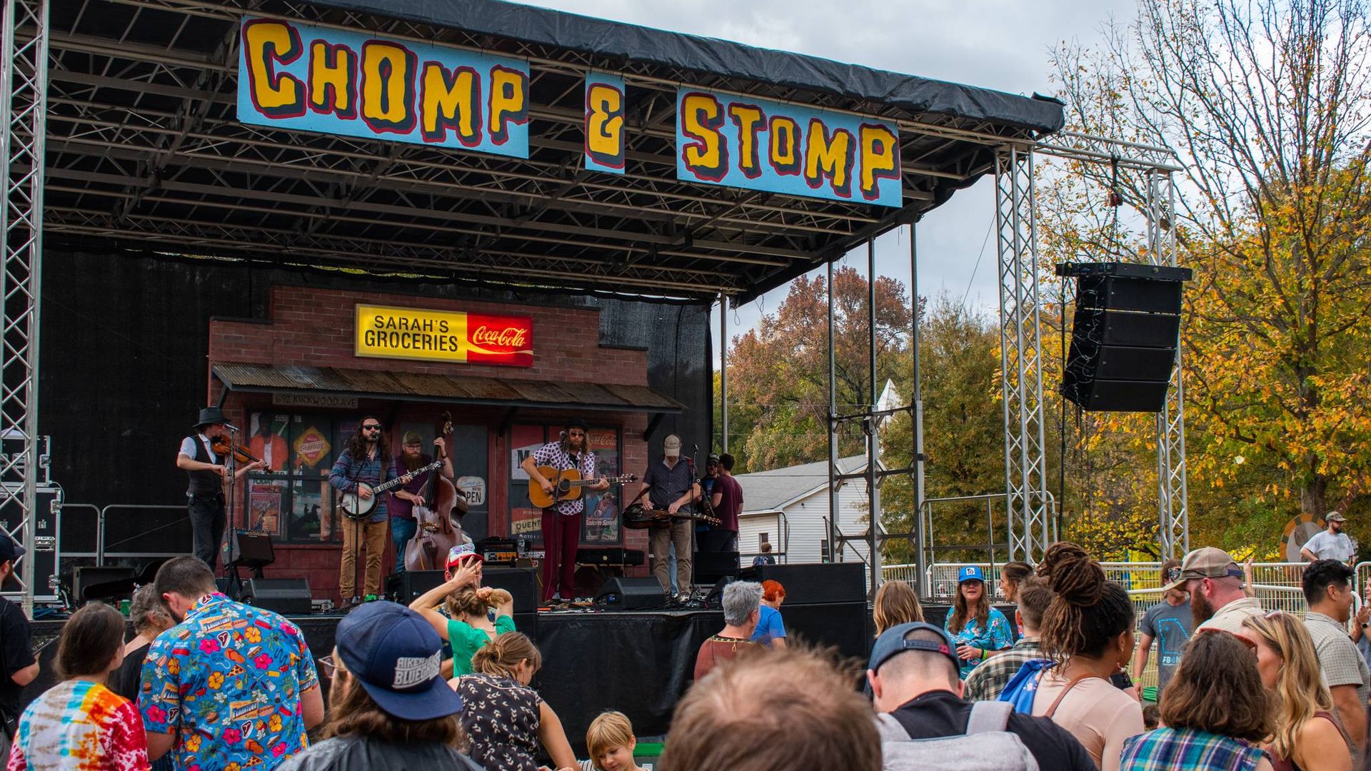 Outdoor stage with band performing under bright signs saying "CHOMP & STOMP" and "SARAH'S GROCERIES Coca-Cola." Audience in colorful clothes watches; trees show autumn leaves.