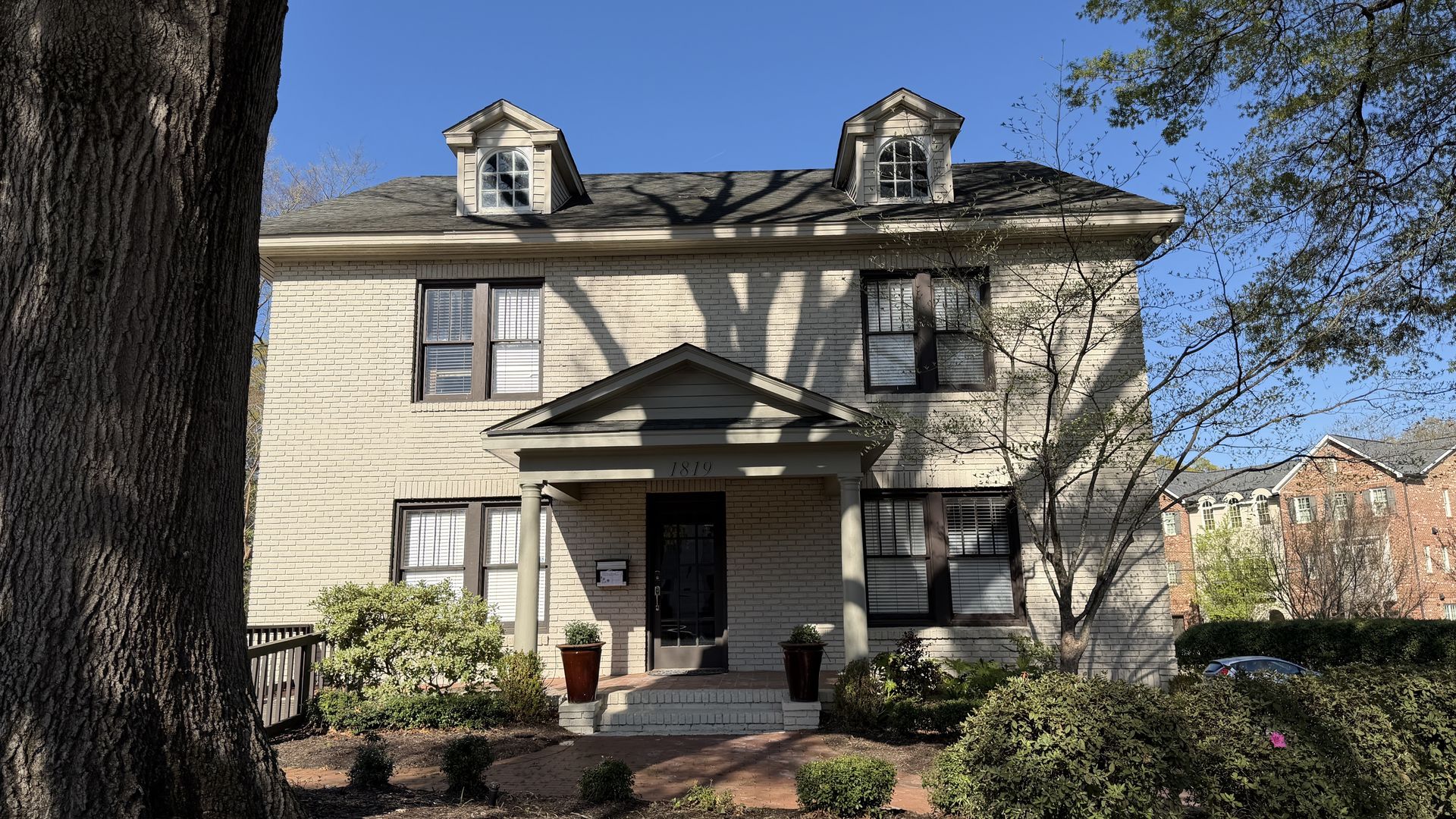 Two-story beige brick house with a centered porch and two dormer windows. A large tree trunk dominates the left foreground; shadows cross the facade. Front steps have two pots, blue sky above.