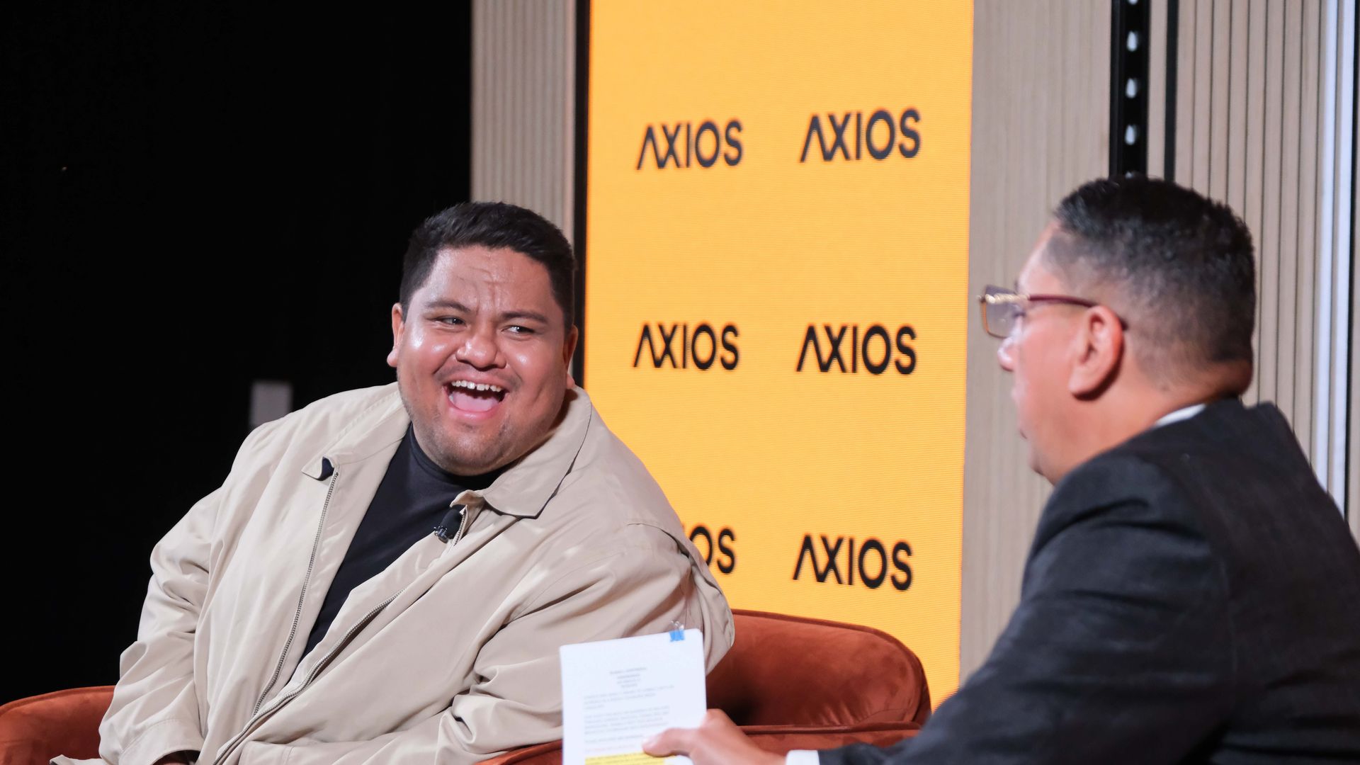 Two men on a stage chat; the man on the left in a beige jacket laughs, the man on the right wears glasses. Behind them is an orange Axios backdrop with repeated logos.