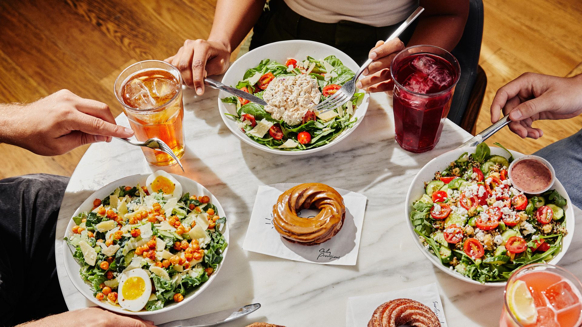 Three people about to eat assorted salads with eggs, cherry tomatoes, cheese, and chickpeas, accompanied by iced drinks and doughnuts on a white marble table.