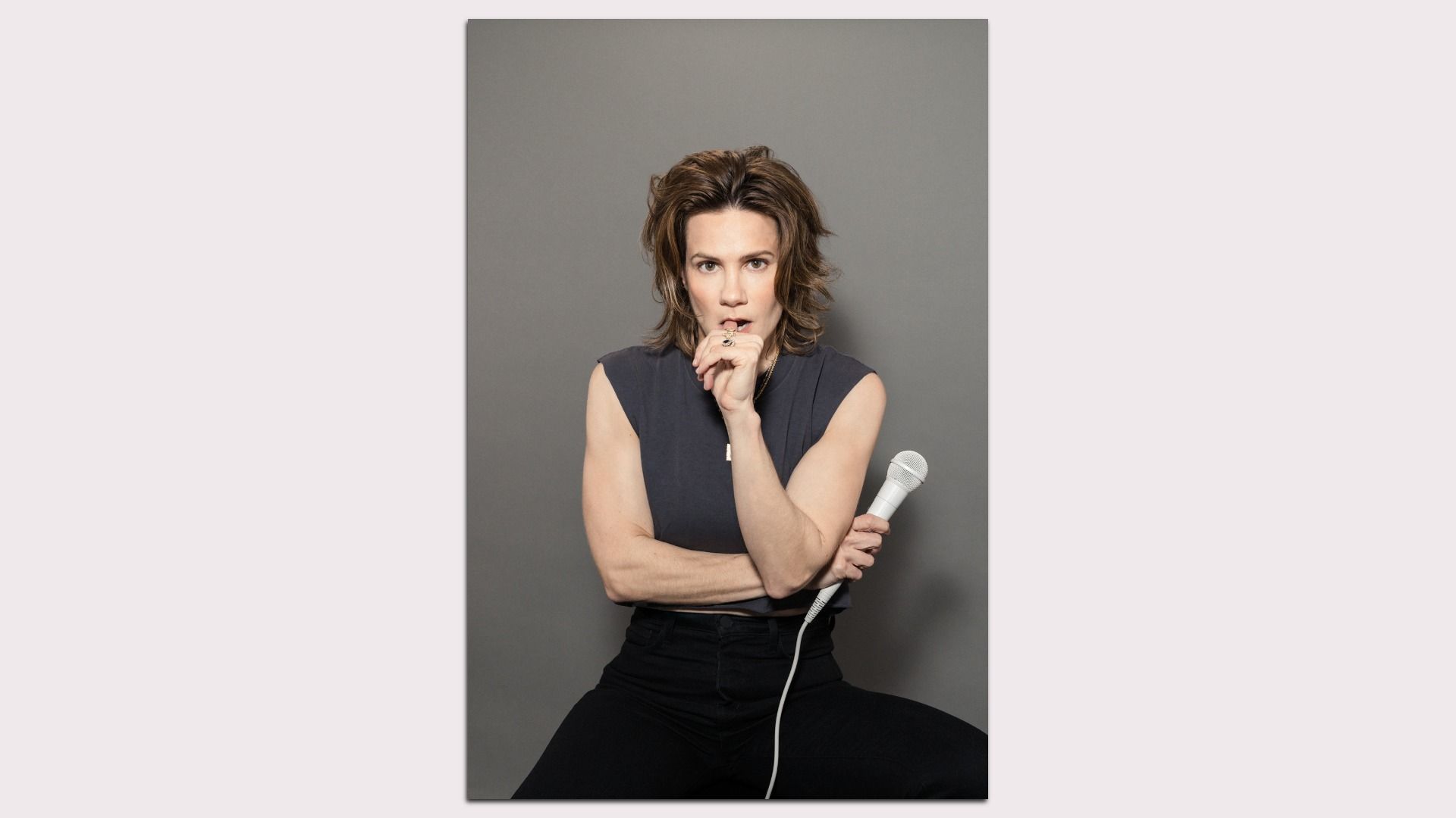 Cameron Esposito with shoulder-length brown hair, wearing a dark sleeveless top and black pants, holding a white microphone and looking thoughtfully at the camera against a gray background.