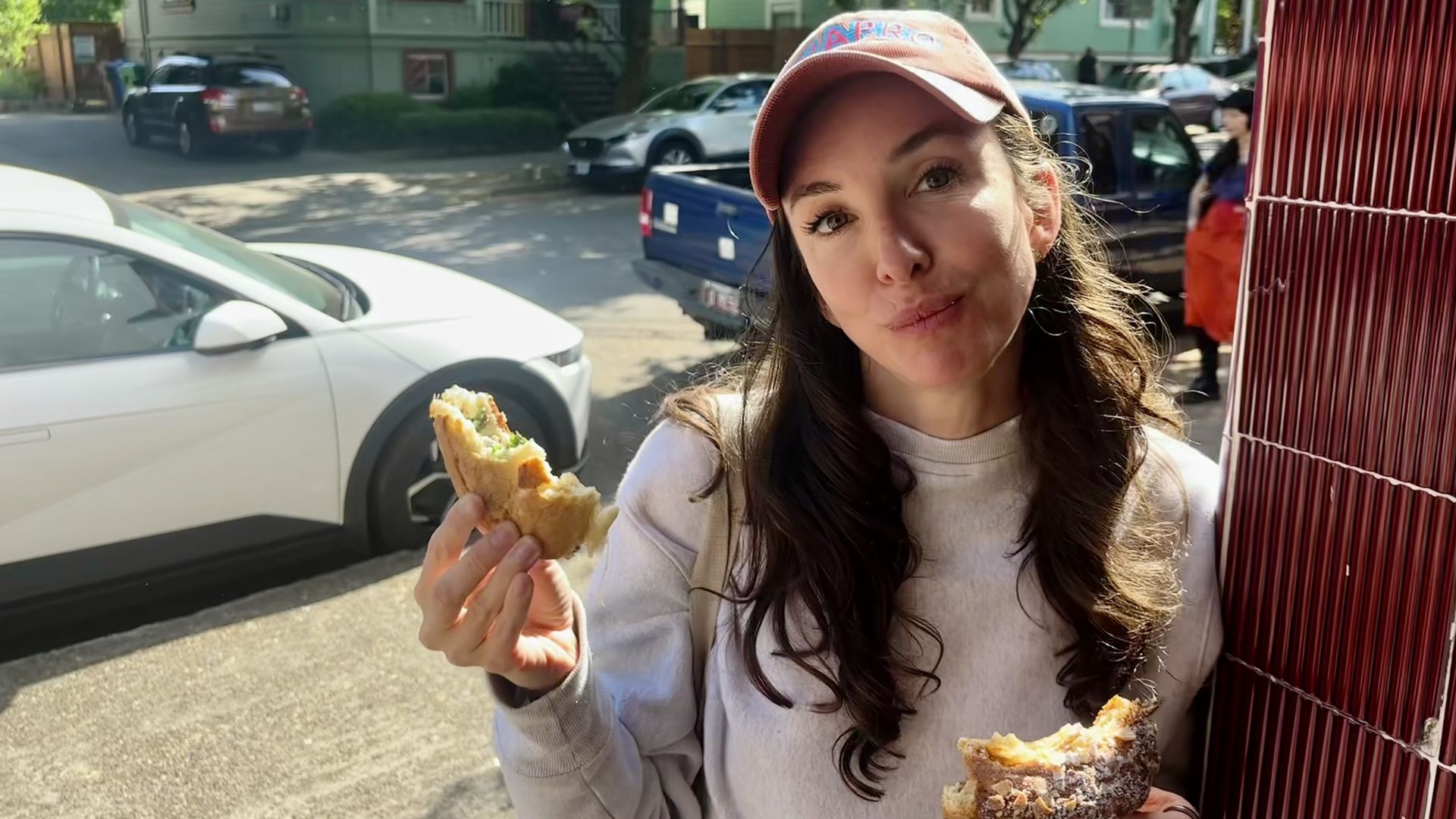 Young woman with long dark hair in a coral-pink cap munches a sandwich outdoors. She holds a bitten crusty roll; street with parked cars, a red bamboo screen, and green buildings in the background.