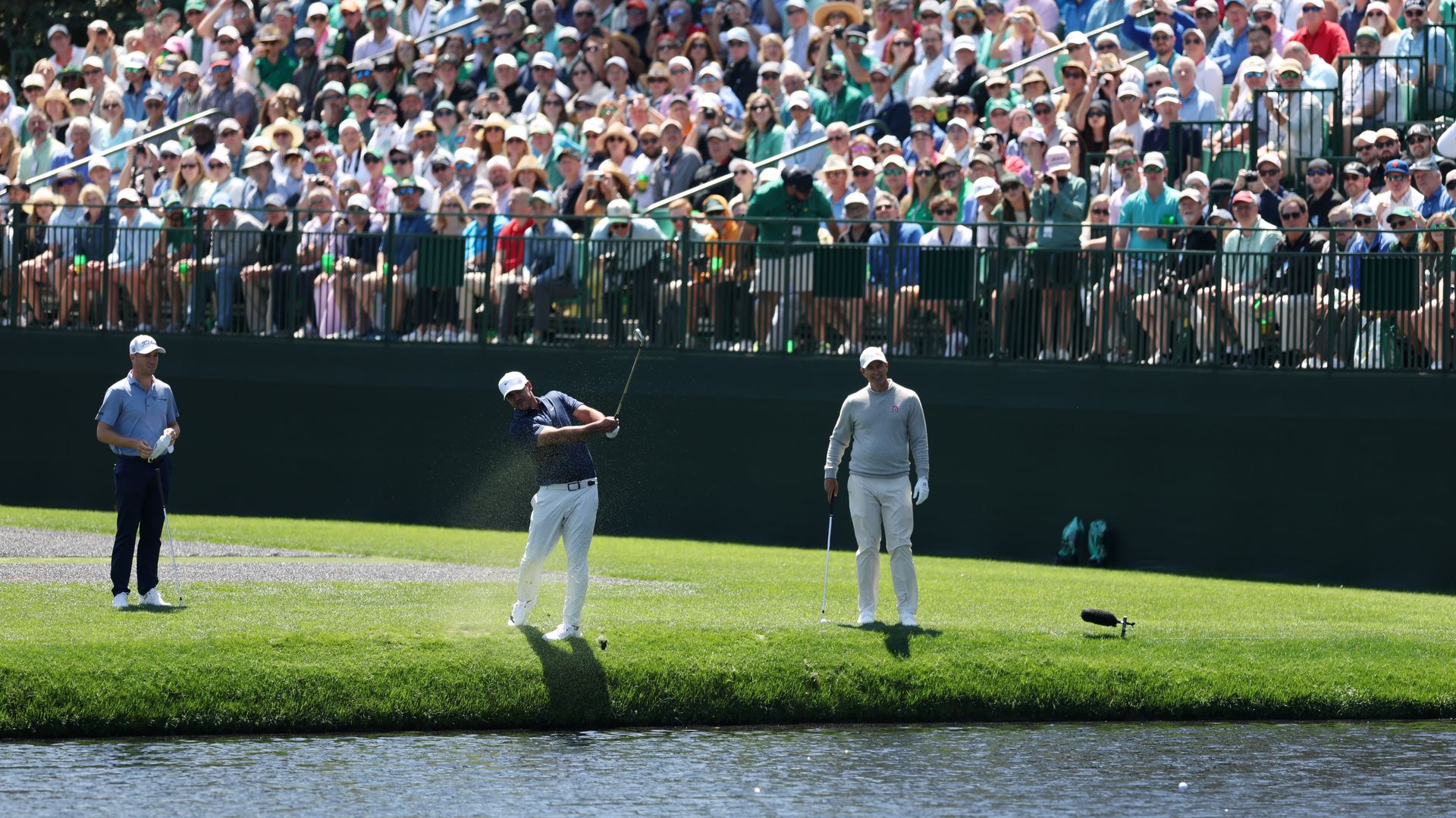 Two men standing and one man swinging a golf club in front of an audience on onlookers. 