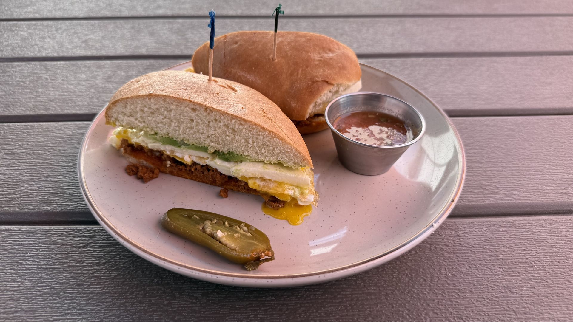 Plate with two sandwich halves containing fried egg, melted cheese, and green sauce, with a metal cup of reddish sauce and a sliced jalapeño.