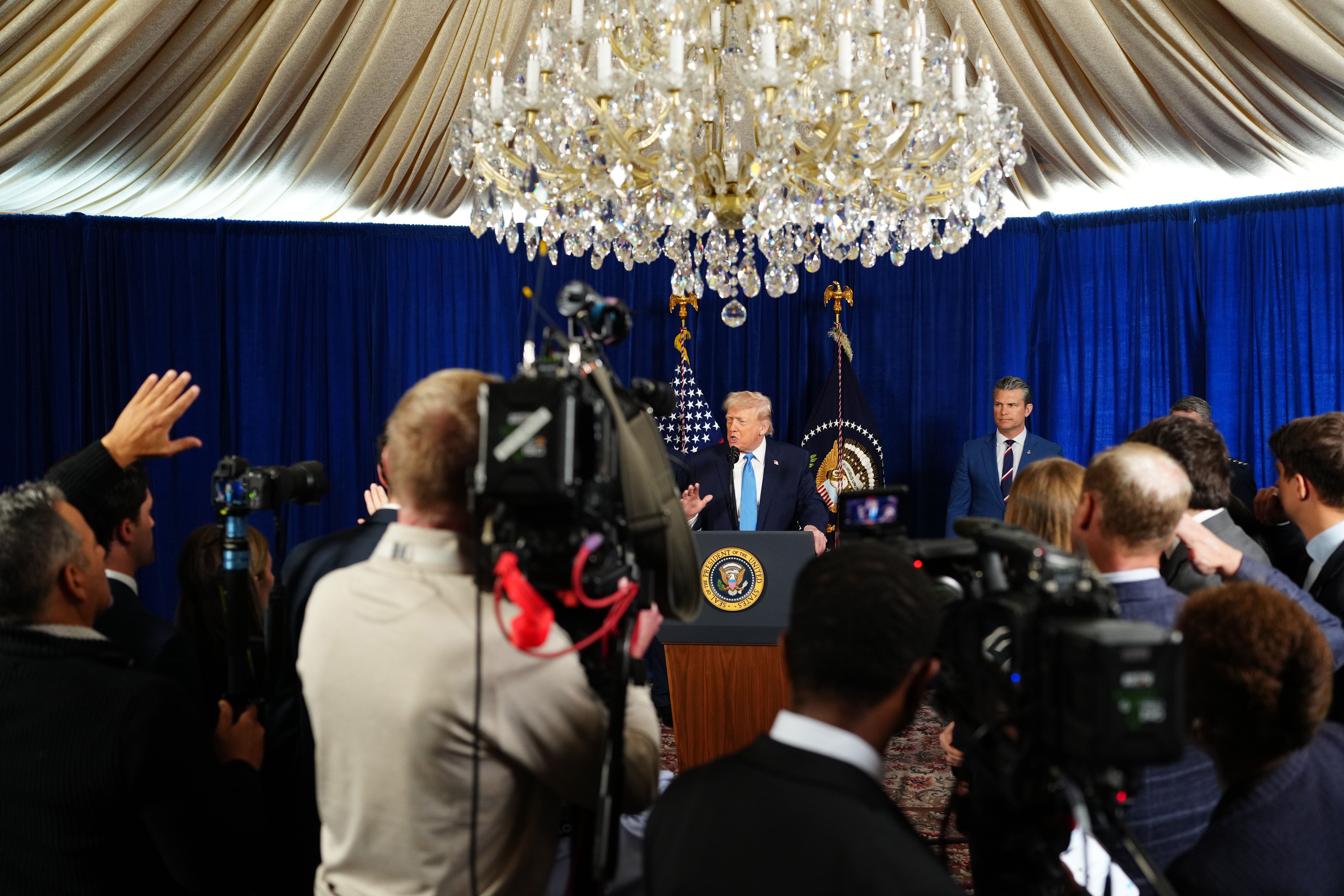 Trump speaking at a podium with the presidential seal, surrounded by cameras, reporters, American flag, and dark blue curtains under a large crystal chandelier.
