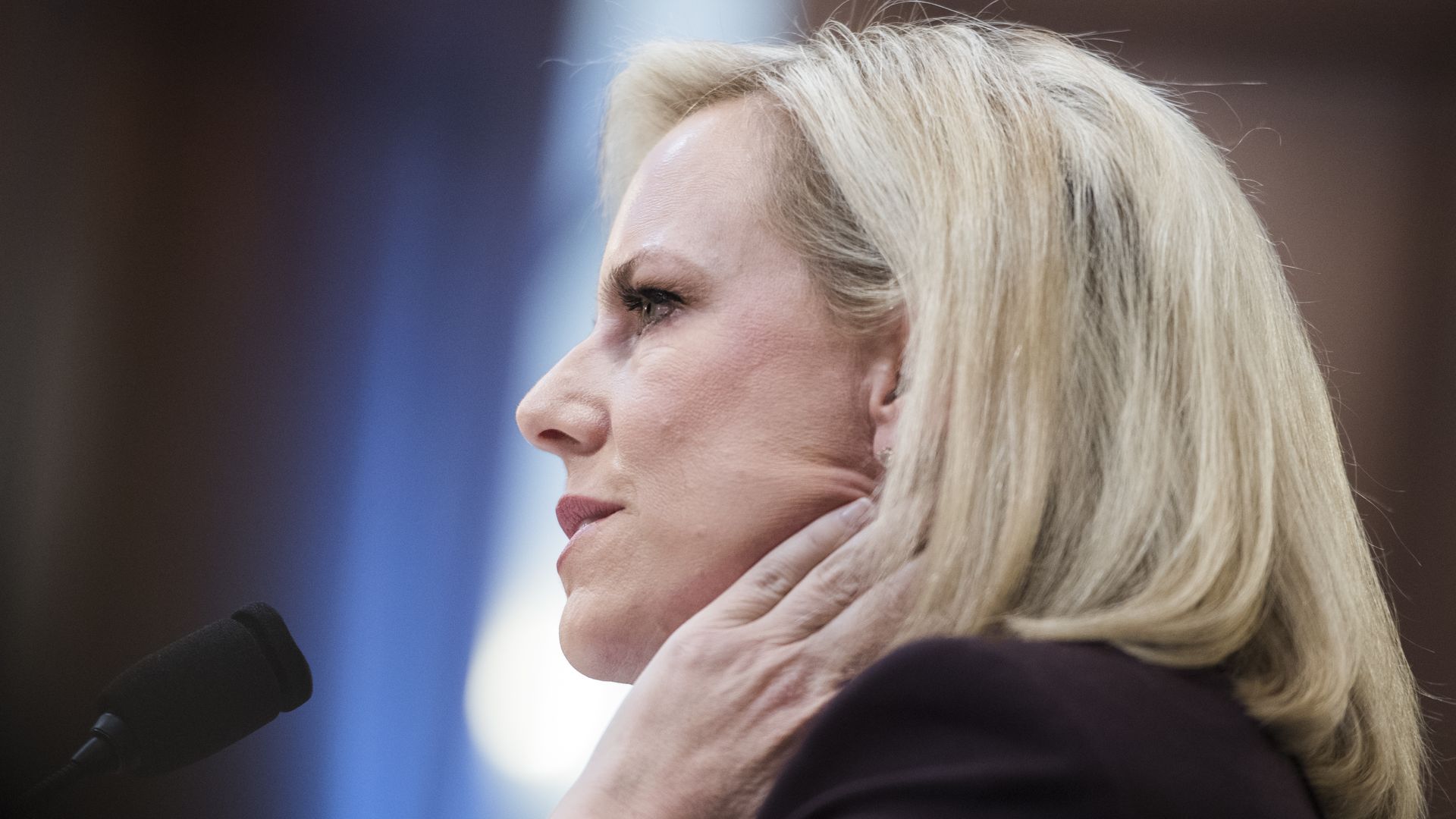 In this image, DHS Secretary Nielsen rubs her neck while sitting and listening in front of a microphone. 