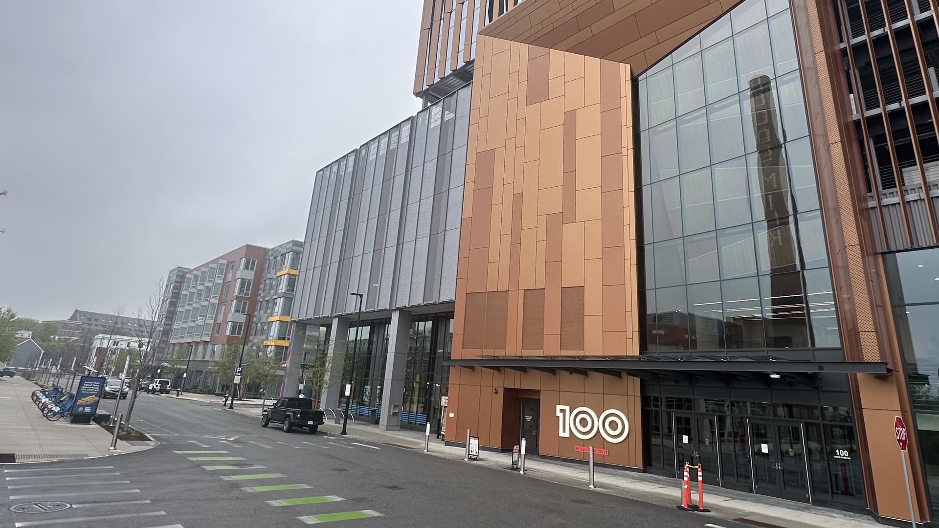 The modern glassy 100 Hood Park building in Charlestown. Down the street are blue bikes and commercial tenants in neighboring buildings, including a nail salon, an apartment leasing office and Tradesman Coffee Shop & Pizza Bar