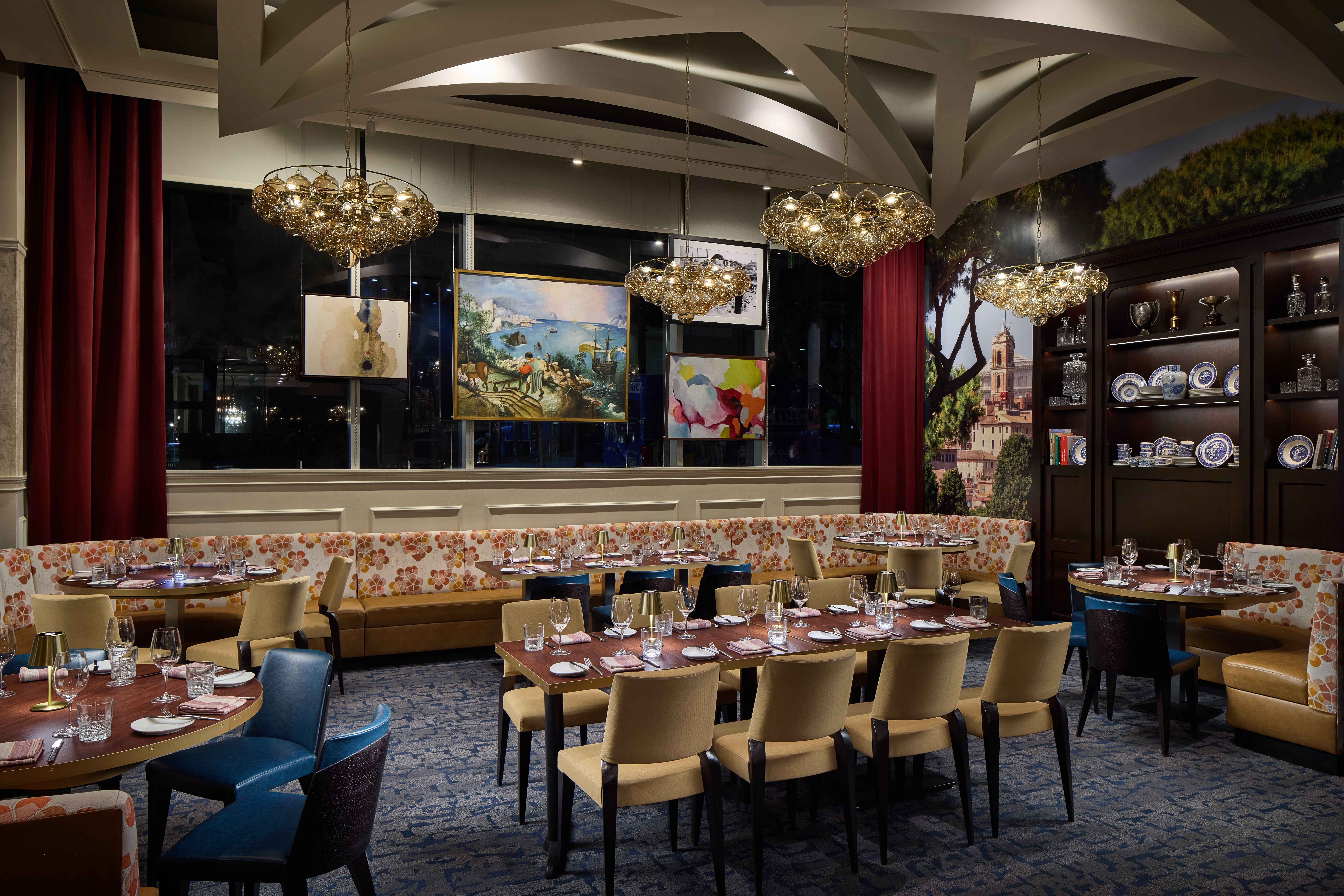 Elegant restaurant interior with round tables set for dining, beige chairs, and a long floral banquette. Glass chandeliers, maroon curtains, dark wood display shelves with blue-and-white china.