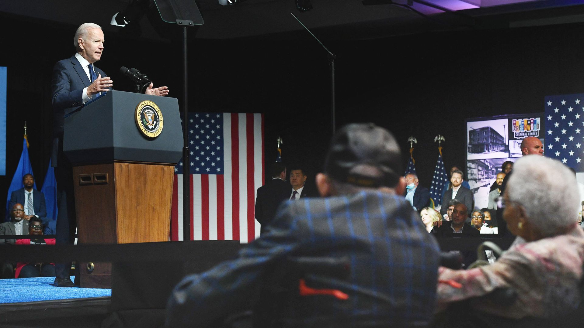 Photo of Biden speaking from a podium while two elderly folks sit and watch