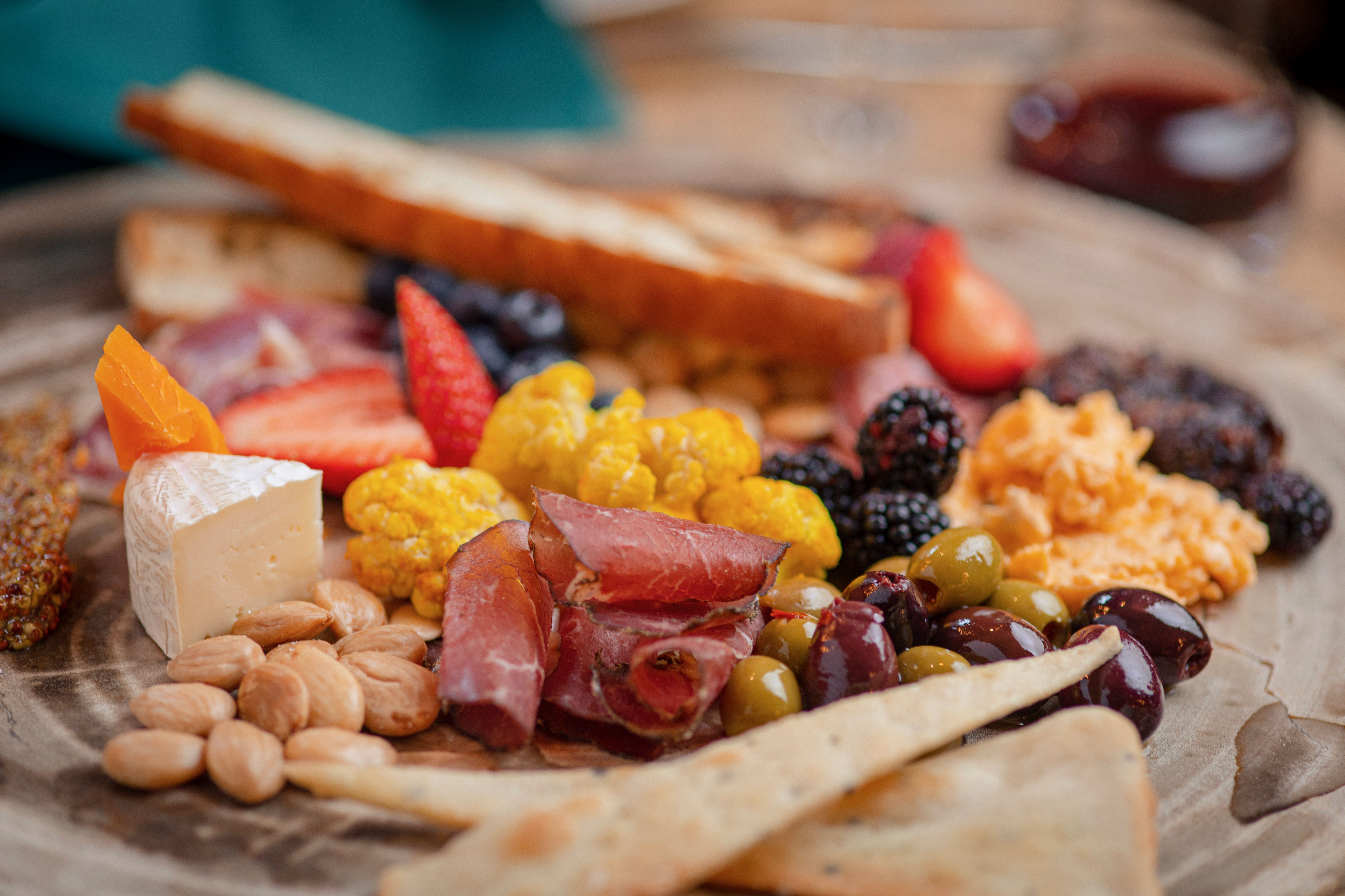 Close-up of a charcuterie board with cheese cubes, almonds, mixed nuts, olives, berries, grapes, sliced meats, and a toasted baguette in the background.