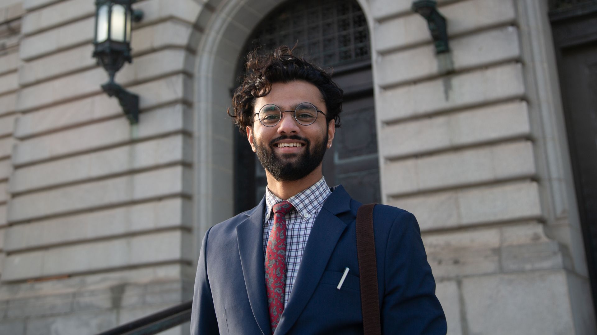 Smiling man with curly hair, beard, and glasses wearing a blue suit, red patterned tie, and checkered shirt stands outside a stone building with arched doorways and lanterns.