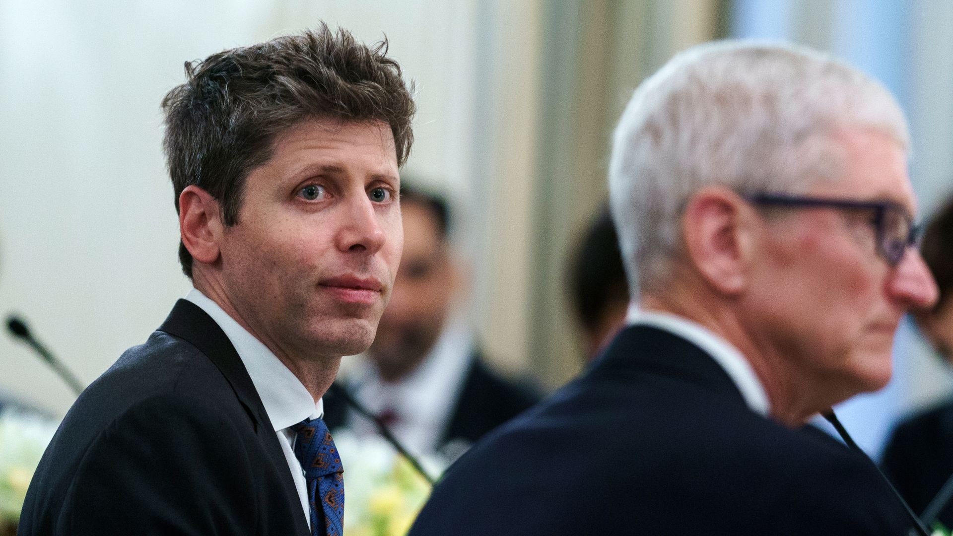 A man in a dark suit and blue patterned tie looks to the left at a formal event, with a man wearing glasses and gray hair blurred in the background.