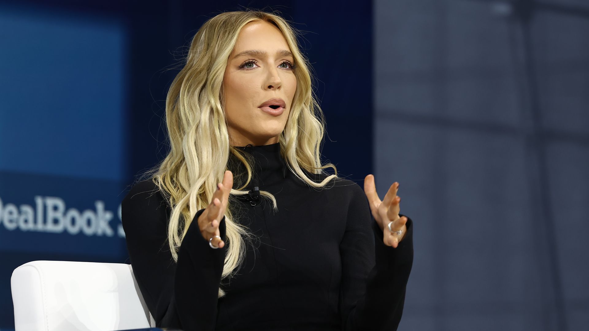 Blonde woman in a black turtleneck dress sits on a white armchair on stage, gesturing with her hands as she speaks. A dark blue backdrop is behind her.