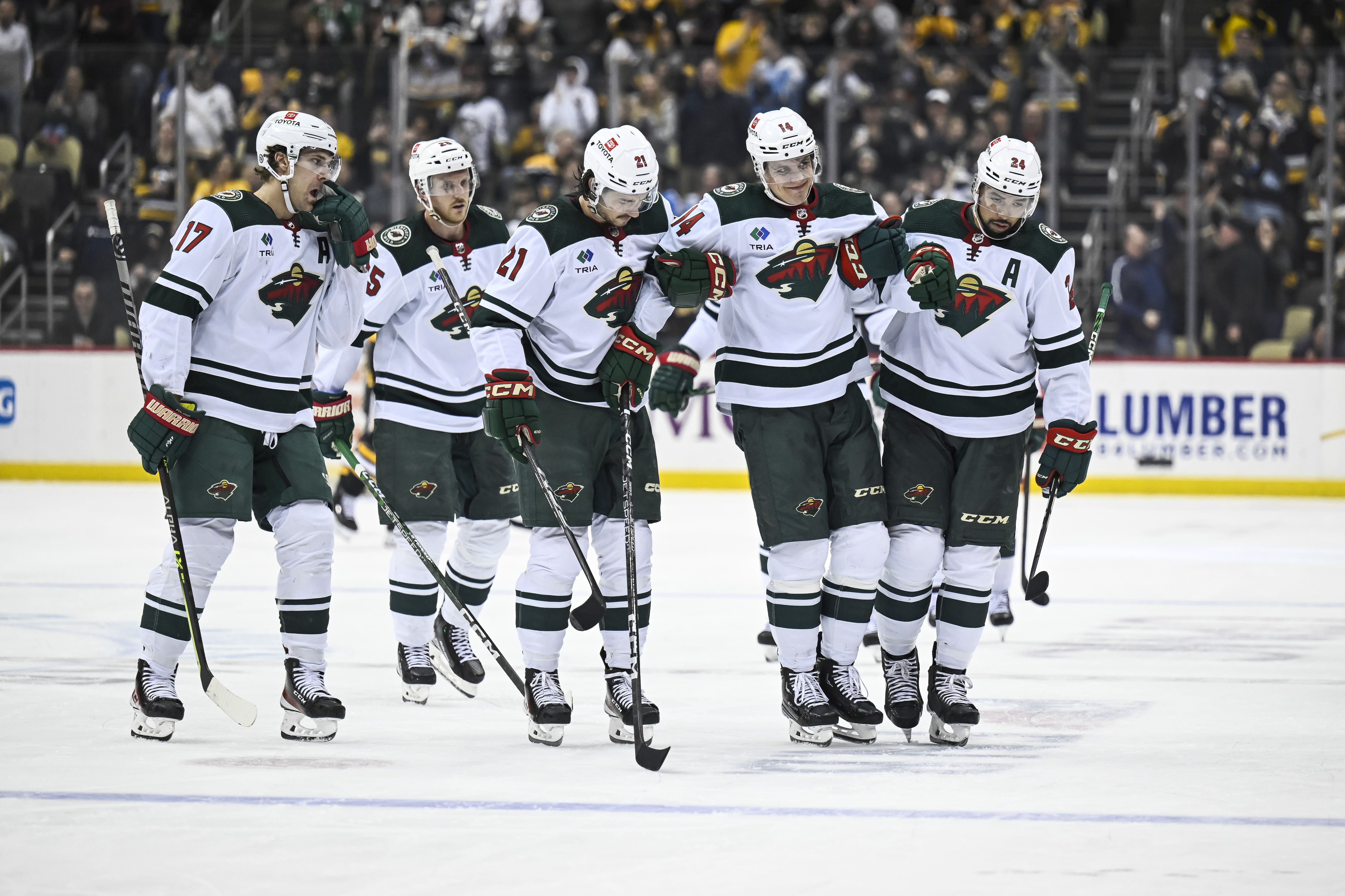 Six ice hockey players in white jerseys with green trim and the Minnesota Wild logo skate off the ice with an injured teammate, sticks raised, holding the injured teammate up by the armpits as he favors one leg.
