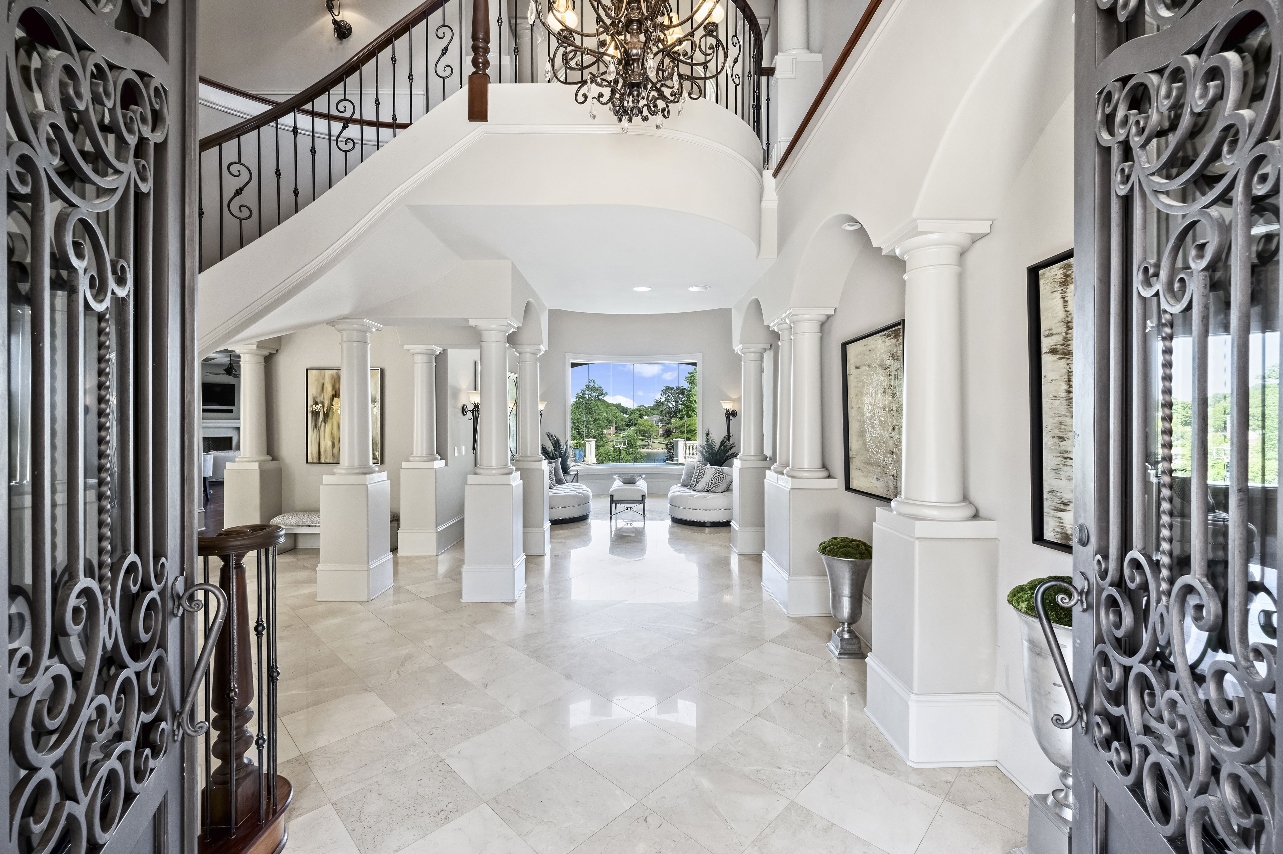 Luxurious foyer with ornate iron gates, curved staircase, chandelier, and white columns. Bright seating area and windows reveal green landscape outside.