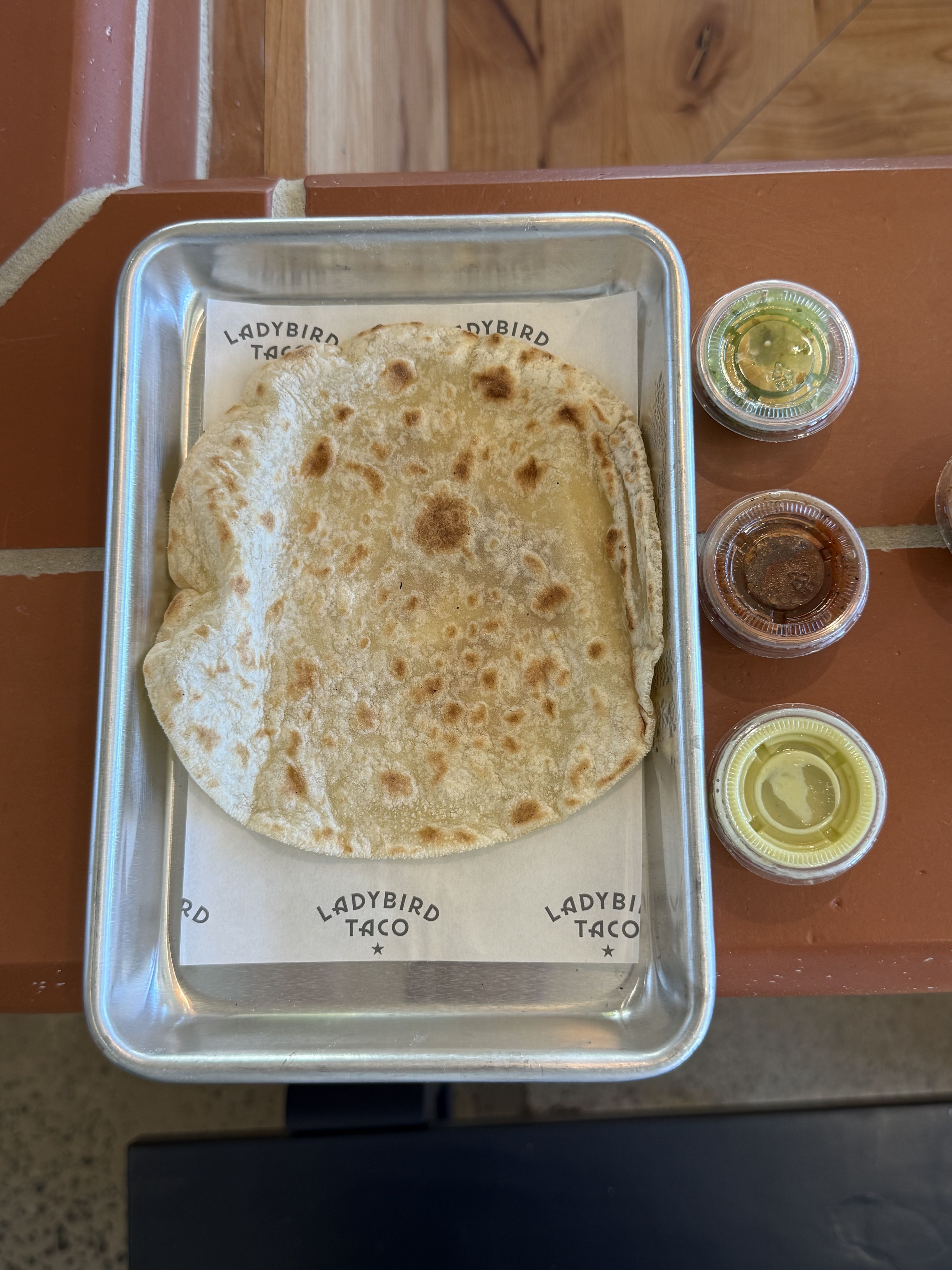 Top-down view of a metal tray with a large floury flatbread, browned spots, on parchment reading LADYBIRD TACO. To the right are small sauce cups in green, brown, and yellow on a wooden table.