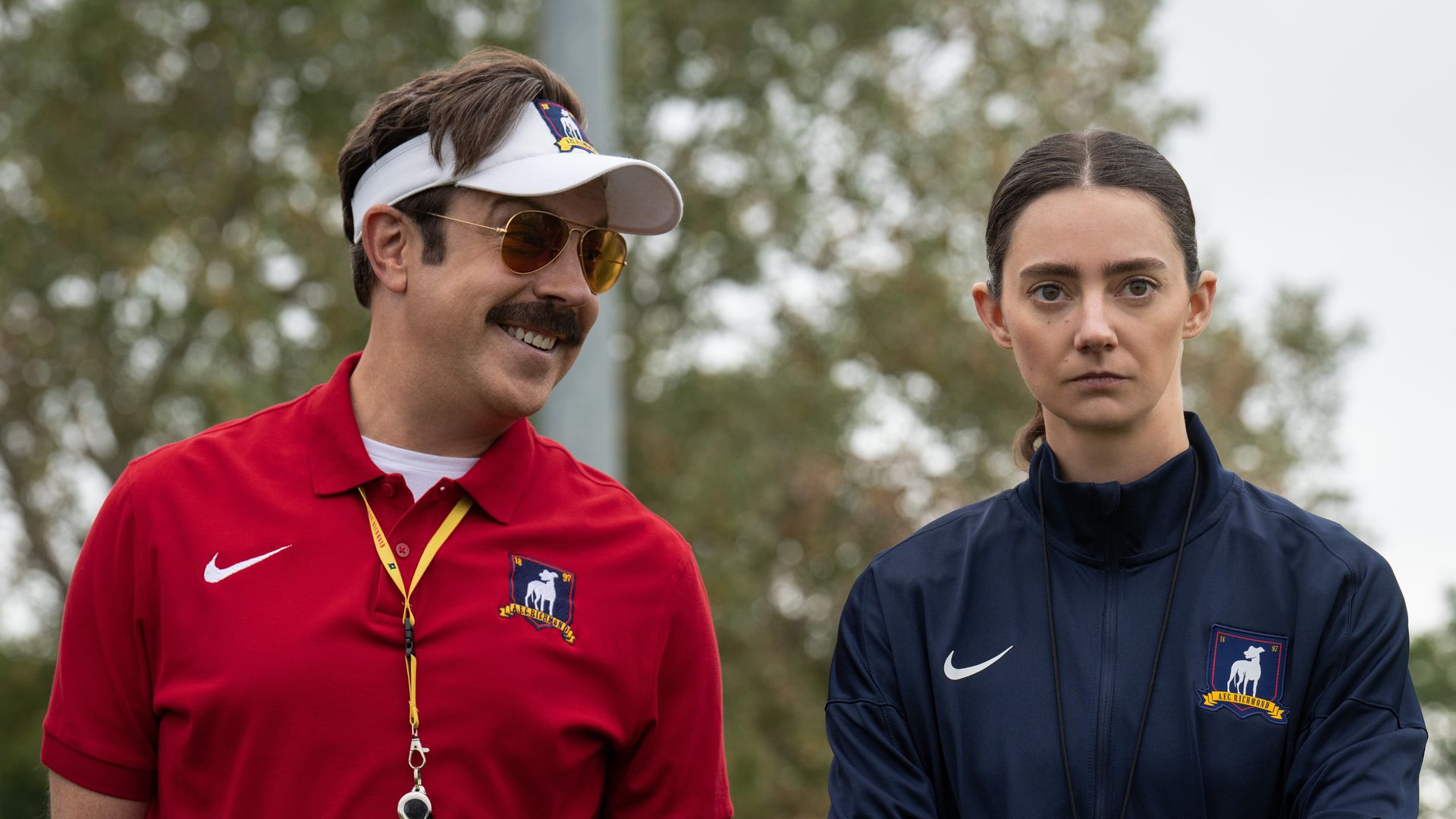 Two adults outdoors: a man in a red polo with a white visor, gold sunglasses, and a lanyard smiles on the left; a stern woman in a navy jacket with a badge stands on the right; blurred trees behind.