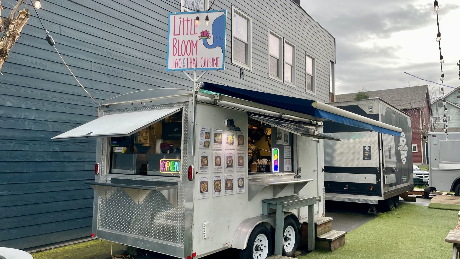 A silver food truck named "Little Bloom Lao and Thai Cuisine" is parked next to a building, displaying a colorful menu and open signs, with string lights overhead and another food truck nearby.