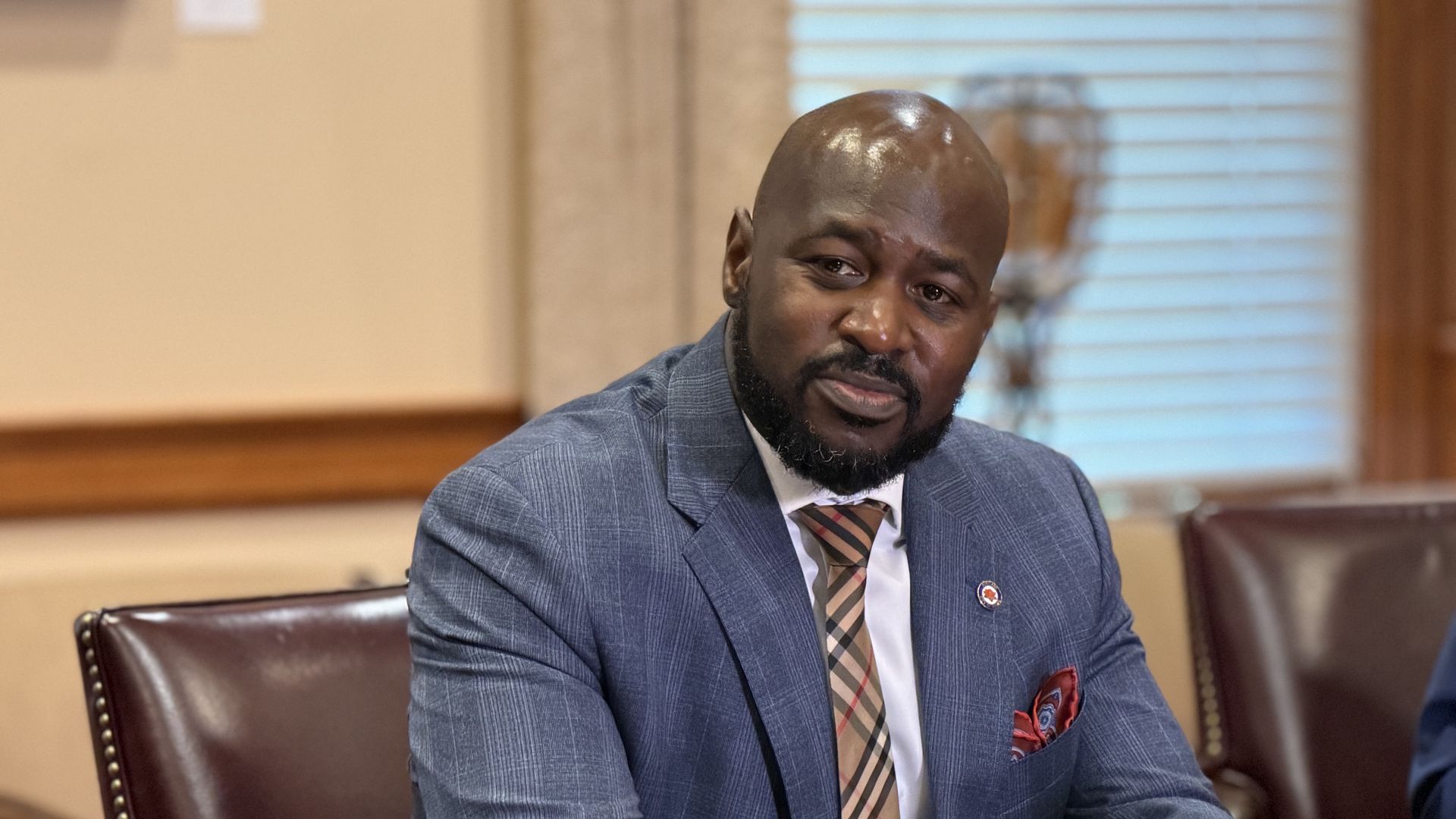 Man in a blue suit with a plaid tie and red pocket square sitting at a wooden table in a room with brown leather chairs and window blinds.