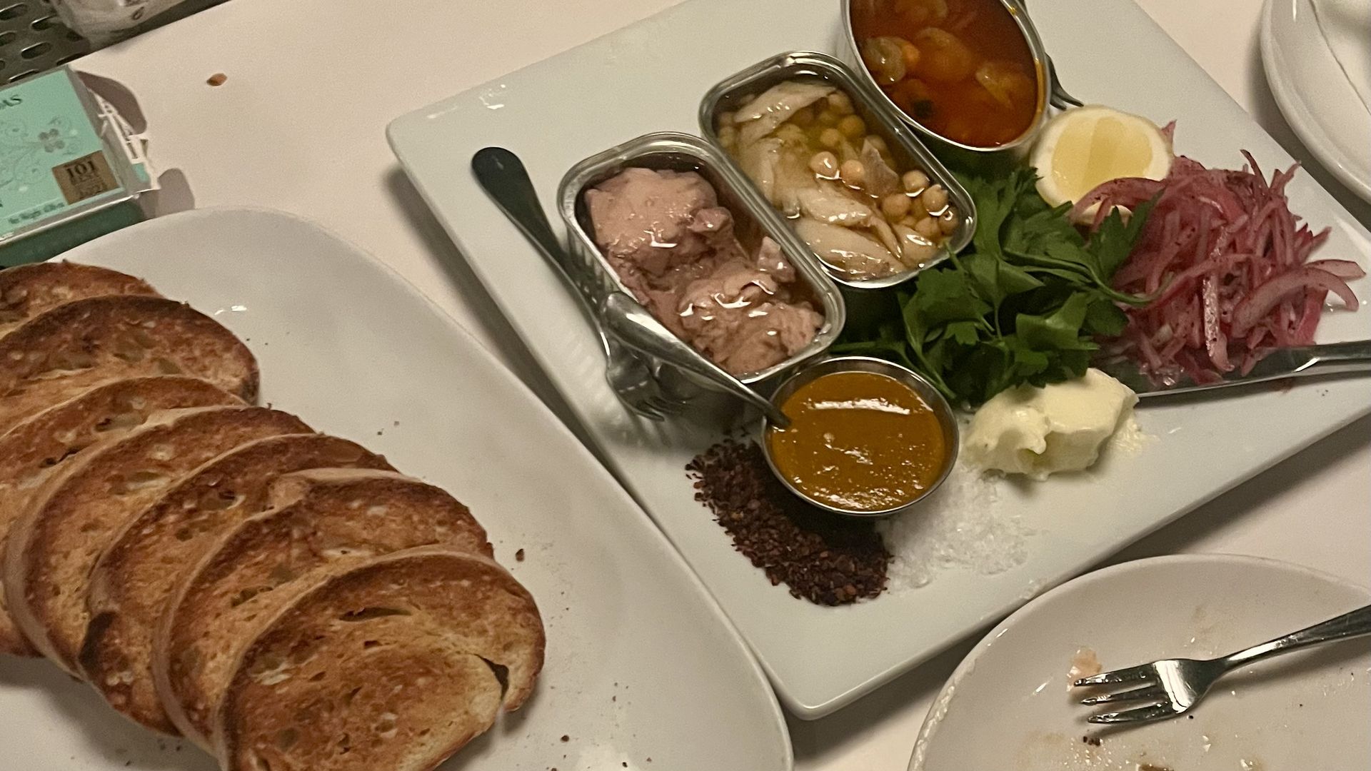 Plates with toasted bread slices and a dish containing pâté, garnishes including onions, parsley, lemon, a brown sauce, and a dollop of butter on a white table.