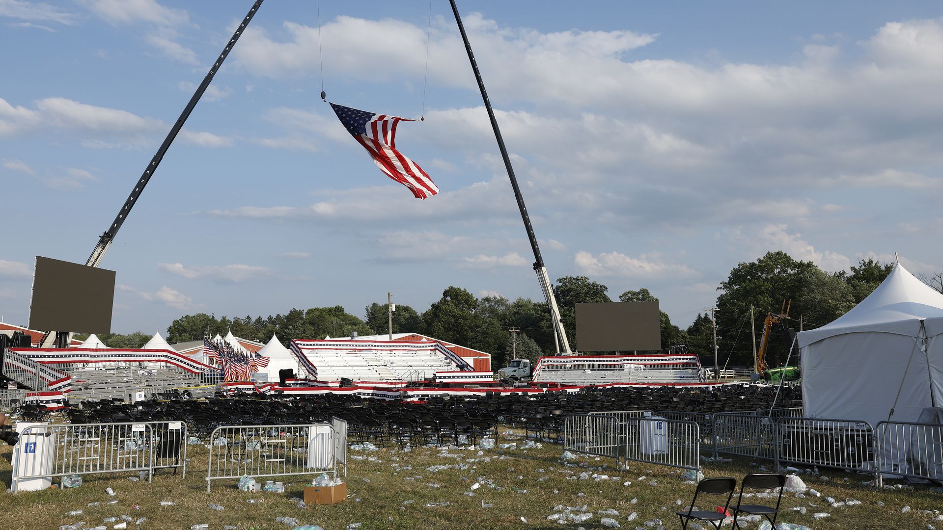 A photo of security railings, tents and crowd litter in an empty rally area with a large U.S. flag suspended between two cranes 