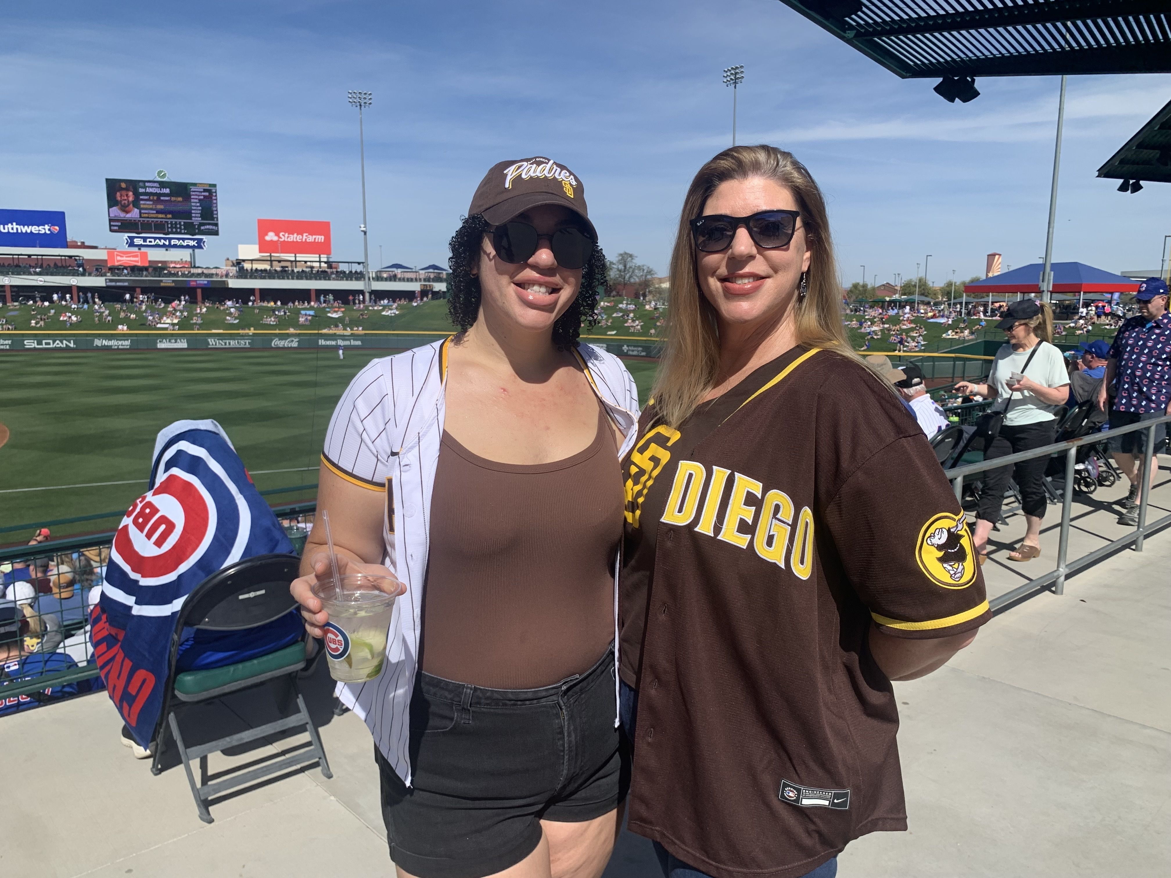 Two women at a sunny baseball park, one wearing a San Diego Padres jersey and cap, the other in a brown top and Padres jersey, with a baseball field and crowd in background.