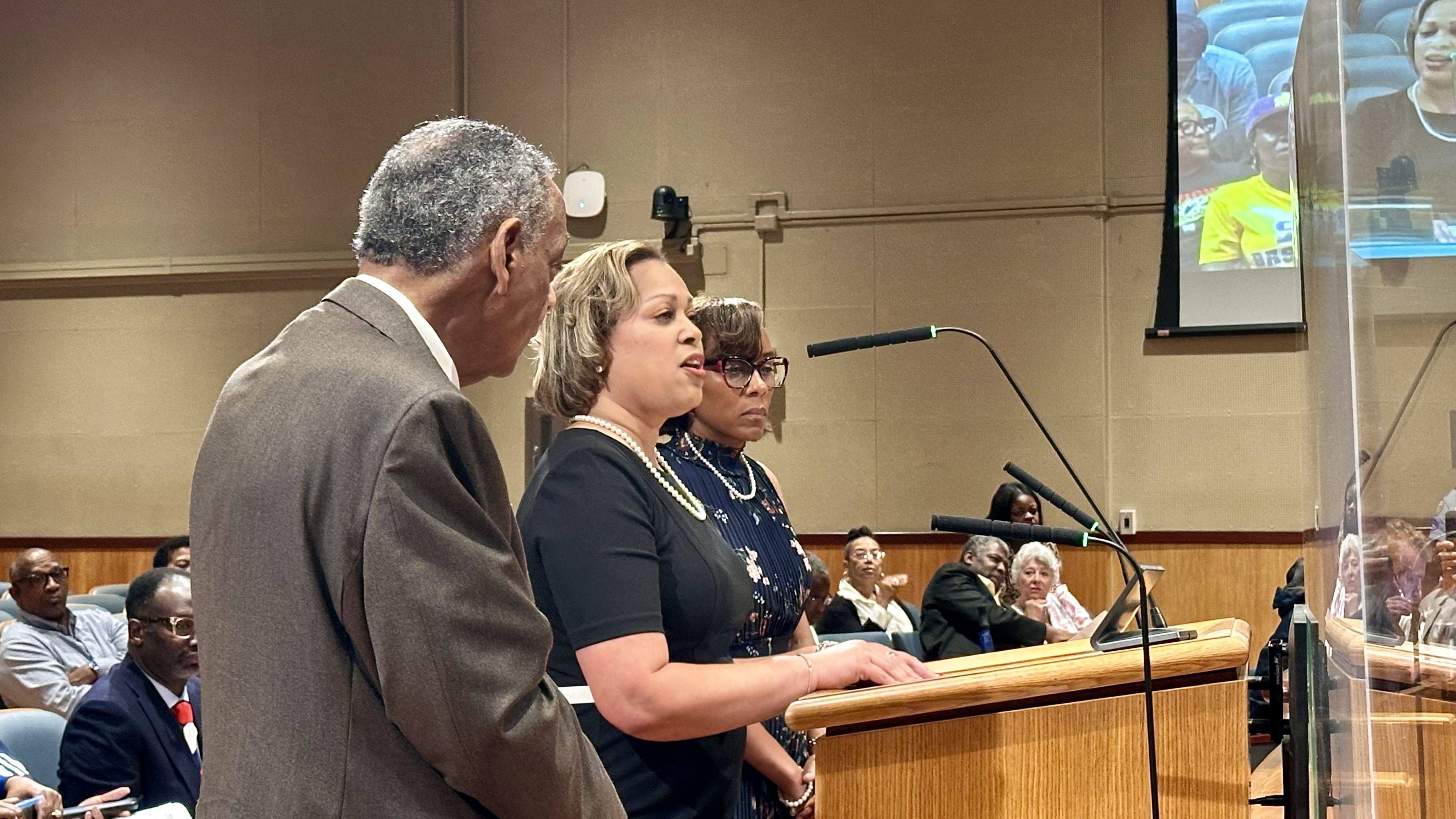 Three people speak at a podium in a conference hall: a woman in black with pearls at center, a man in a suit to the left, a second woman behind; audience seated.