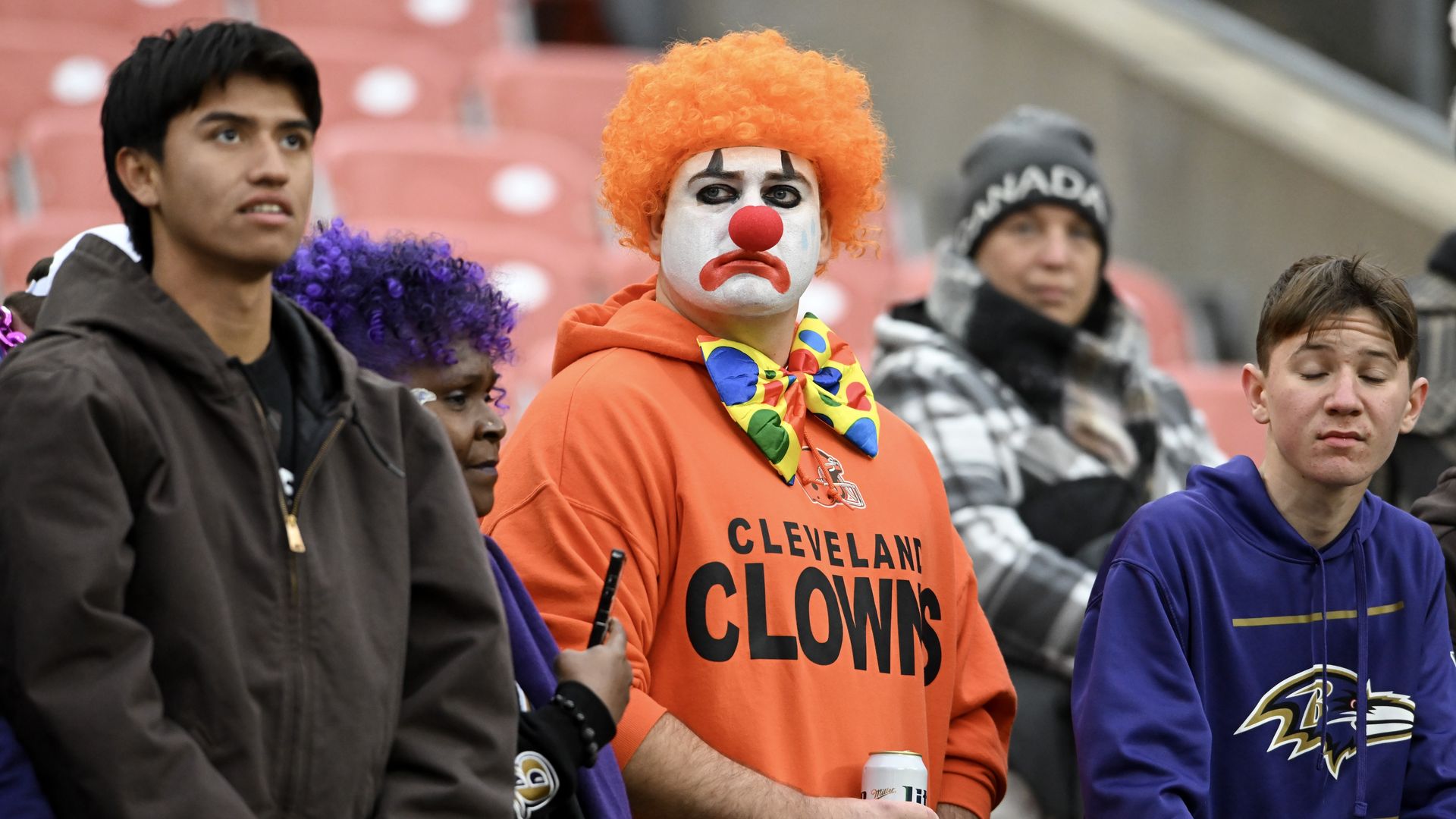 A group of sports fans including a man dressed as a sad clown in an orange hoodie labeled "Cleveland Clowns" with a red nose, orange wig, and colorful bow tie, holding a can.