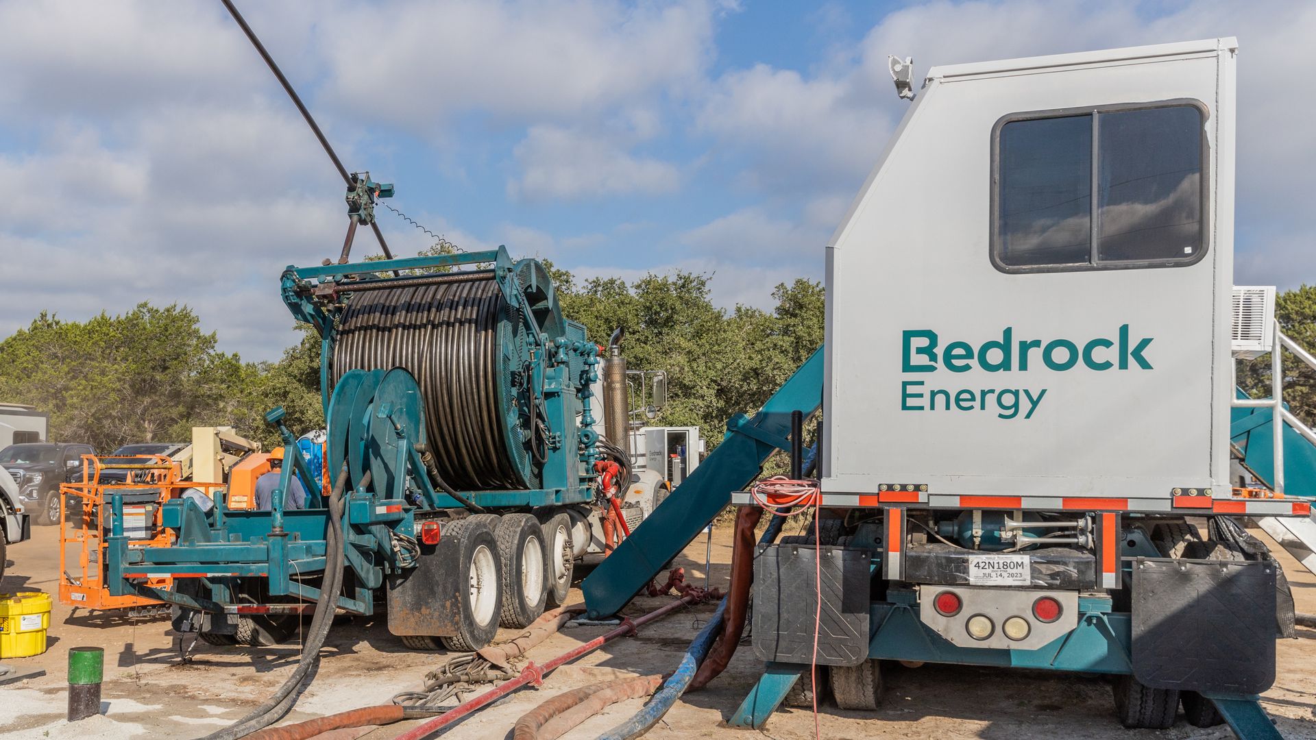 A photo of giant steel cable wrapped around a spool on the back of a green truck, next two another truck with what looks like a generator on the back.
