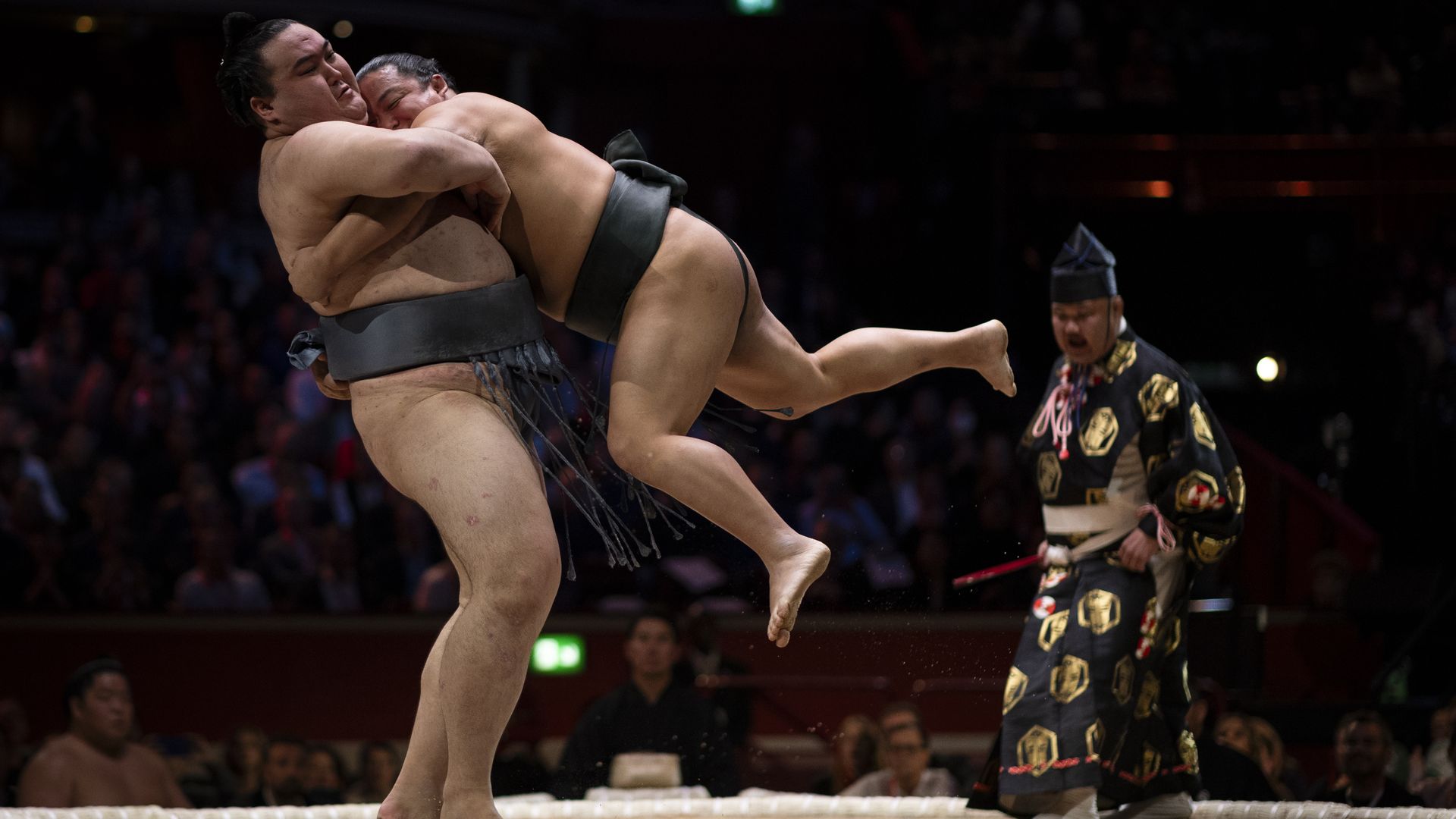 Two sumo wrestlers grapple in the ring, one lifting the other as they clash. A ceremonial official in a patterned robe watches nearby, with a dark arena and audience in the background.