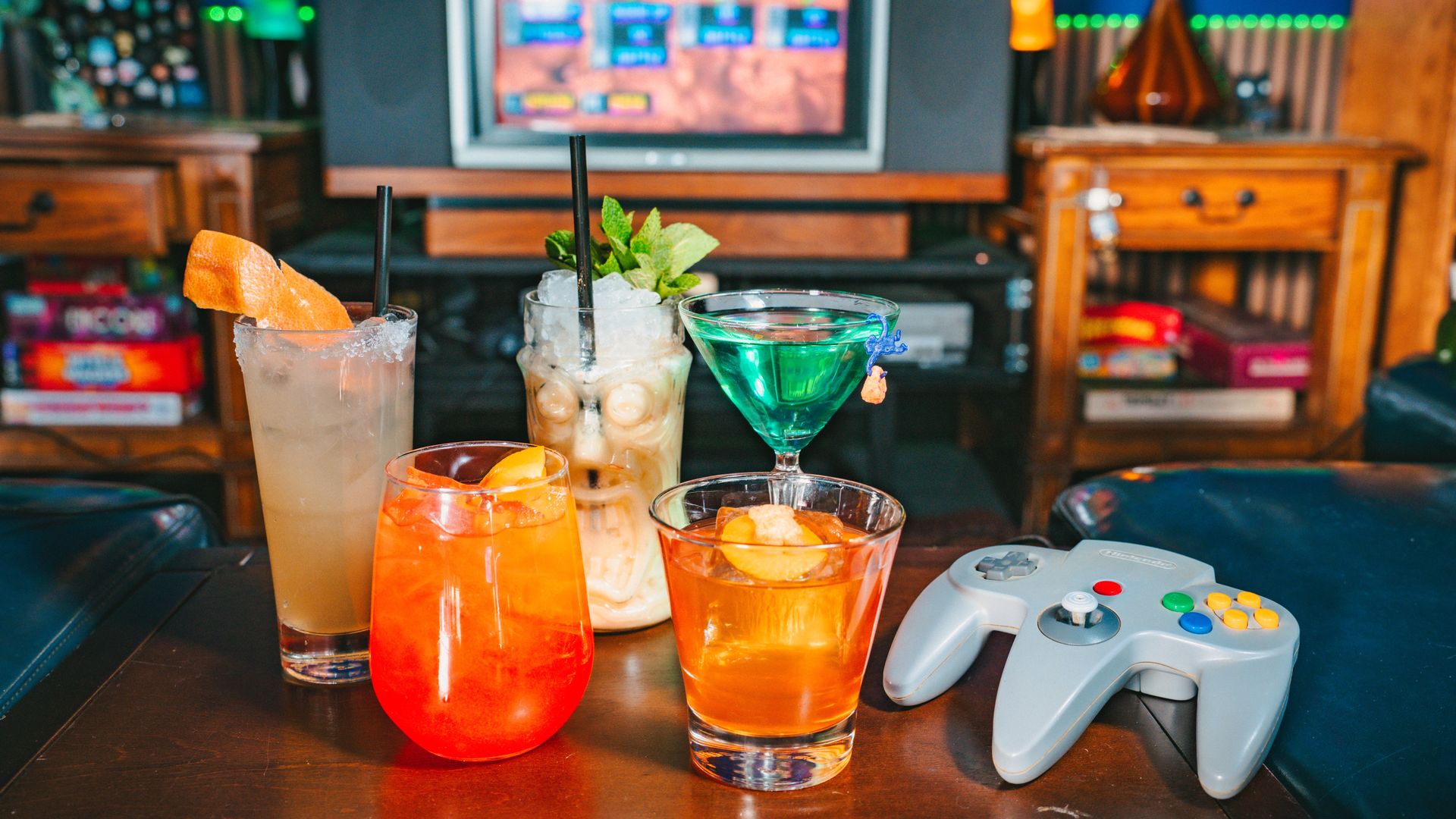 Cocktails on a table: pale long drink with orange wedge, mint mojito, skull glass, green martini, and amber drink; a gray game controller sits nearby with a TV and game shelves in the background.