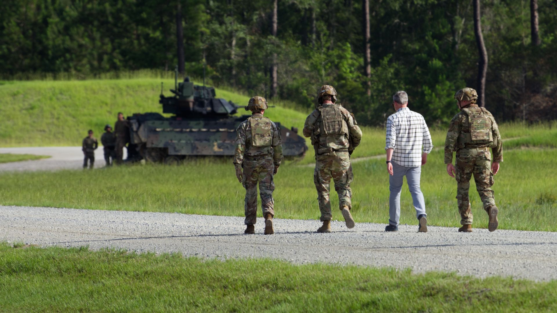 Four men walk down a dirt path. Three are in Army uniforms. At the far end is a large combat vehicle. The surrounding grass and trees are verdant.