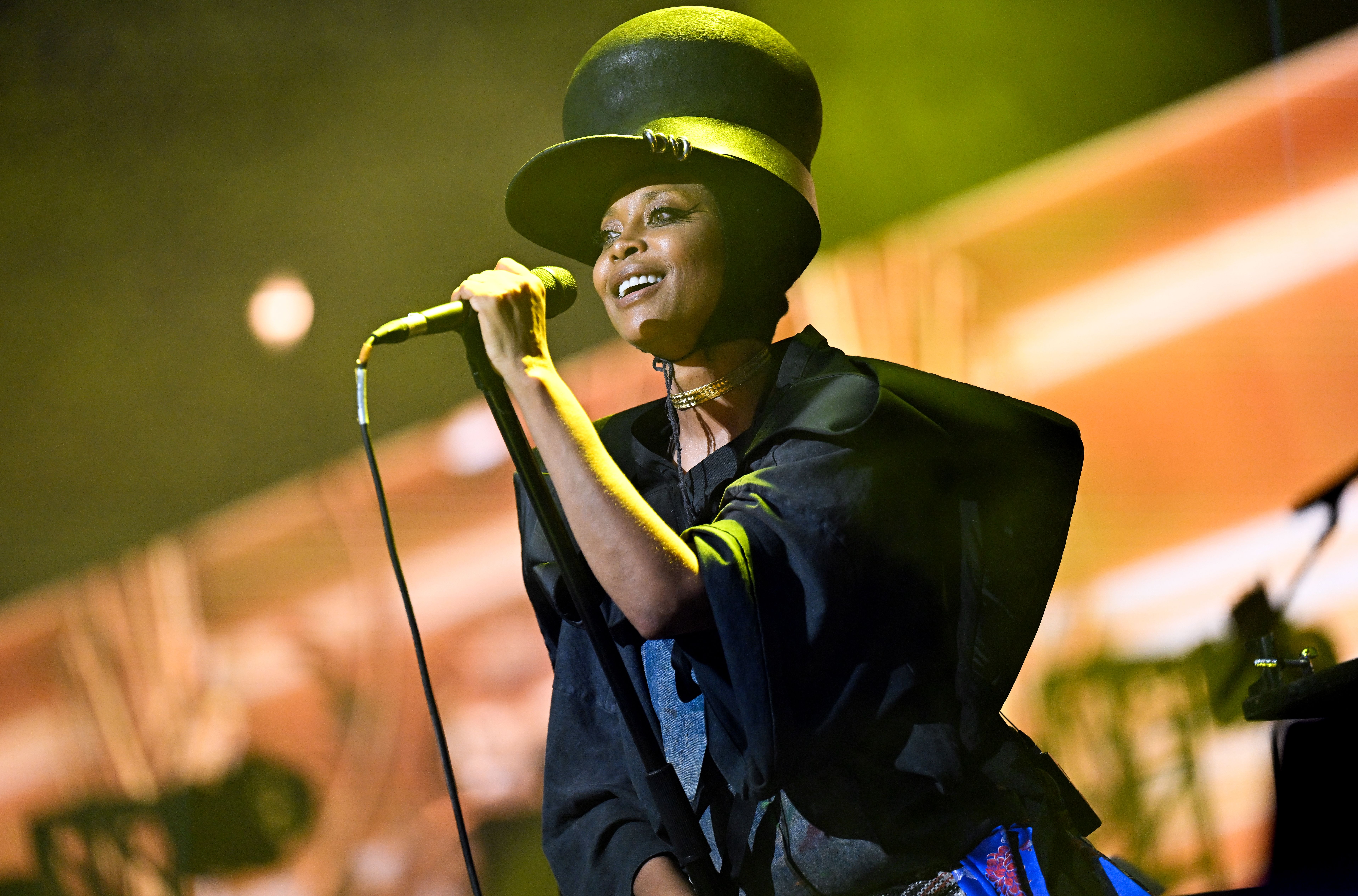 Singer on stage wearing a large black top hat and black outfit, holding a microphone, bathed in yellow and orange stage lights with blurred background.