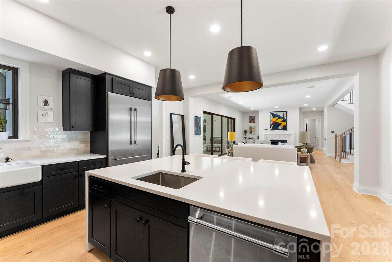Open kitchen with black cabinets, a large stainless refrigerator, and a white quartz island with an undermount sink and black faucet. Two black pendant lights above; living area with colorful art.