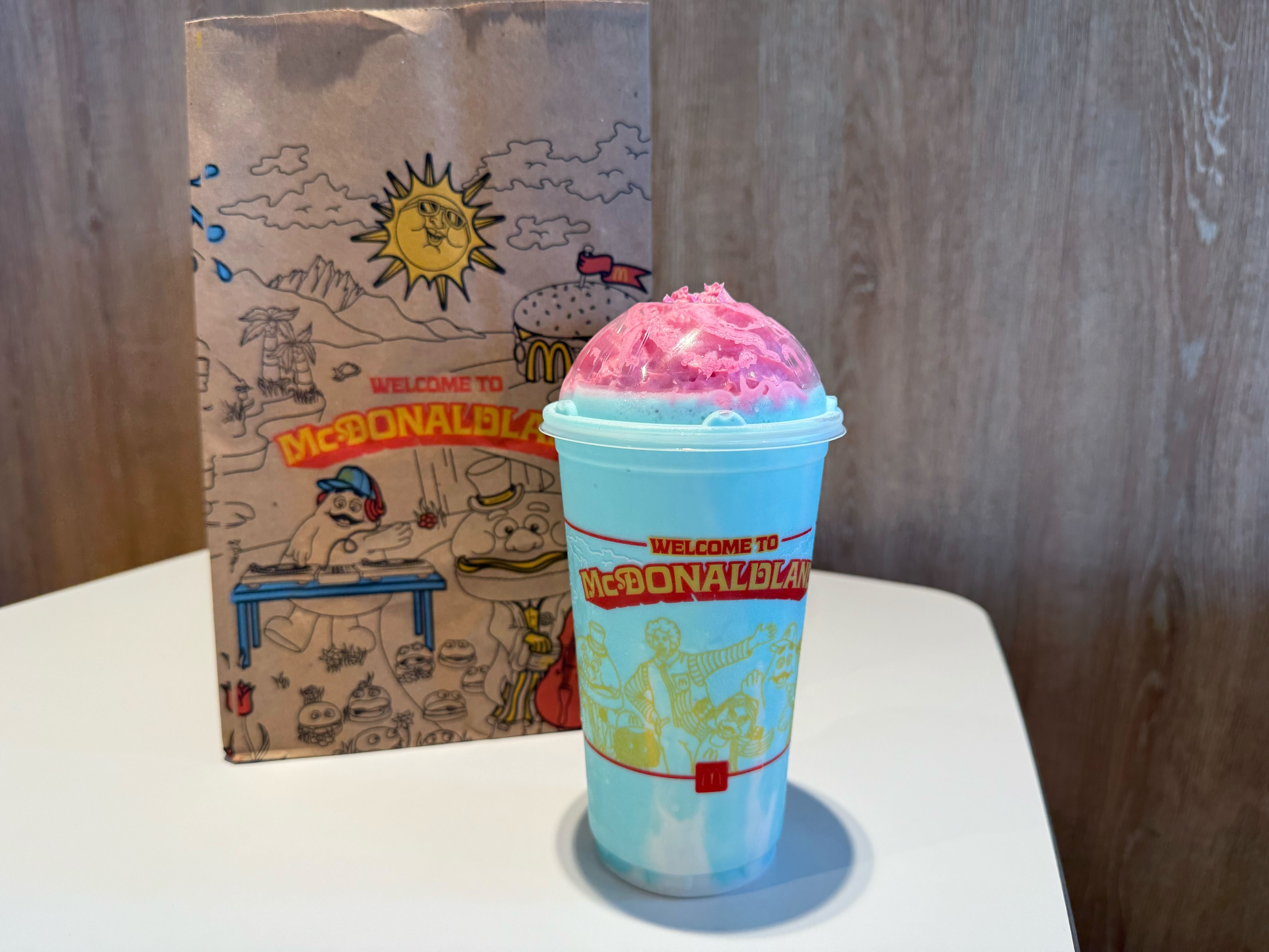 A blue McDonald's shake with bright pink whipped cream in a cup with "Welcome to McDonaldland" design, placed on a white table with a brown McDonald's paper bag featuring cartoon characters in the background.