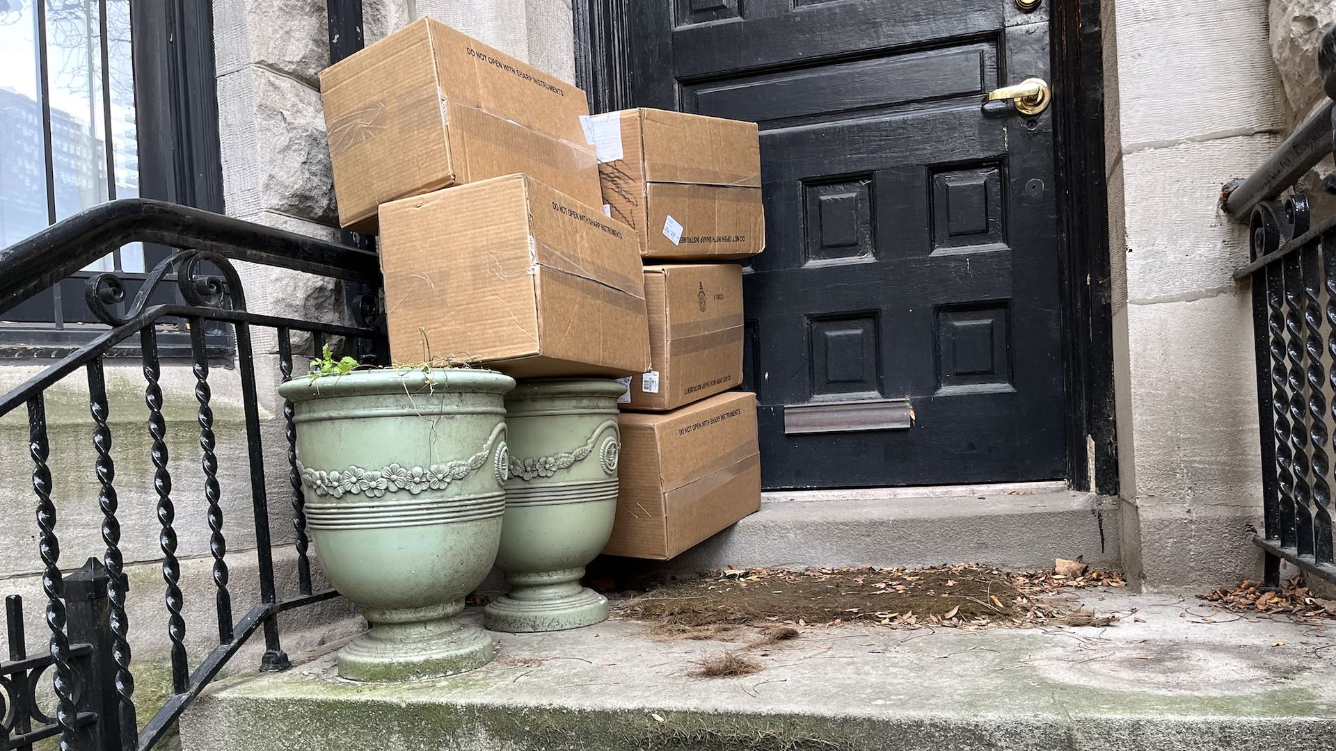 Four cardboard boxes stacked in the corner of a front porch on top of green planters in front of a black front door.
