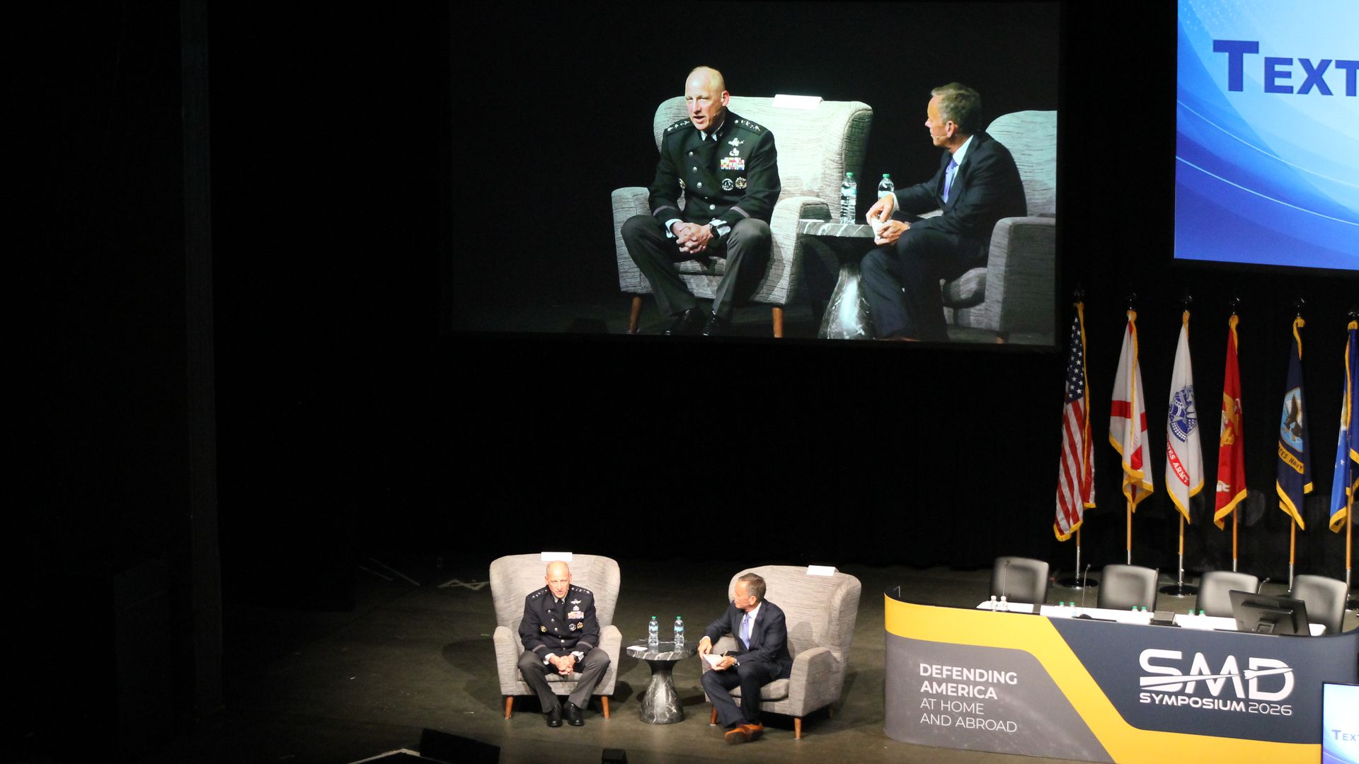 Two men sit on stage in gray chairs; one in a bald military dress uniform, the other in a suit. A large screen shows them, with flags and a blue display to the right.